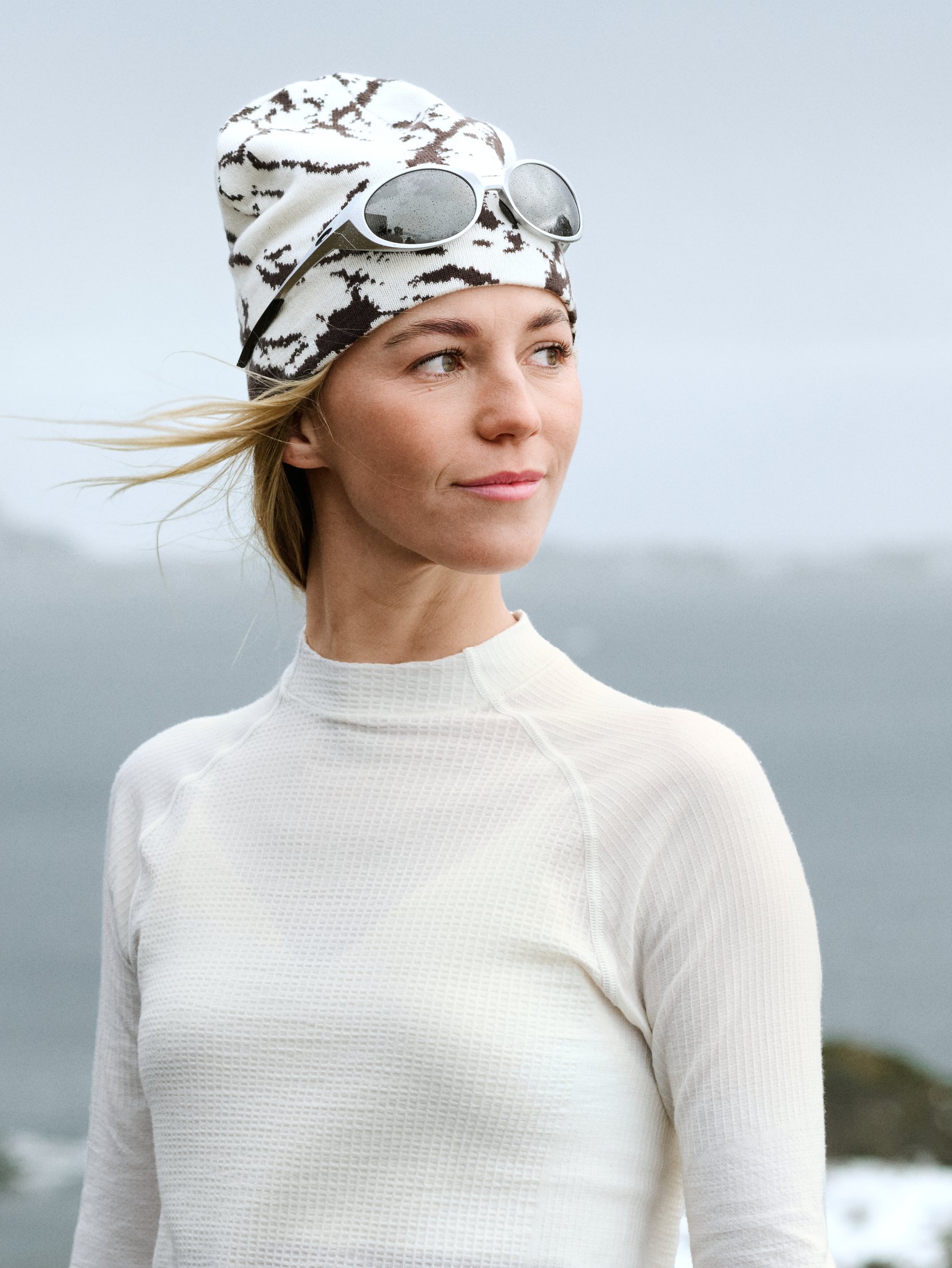 Blonde Frau, die nach rechts schaut, trägt ein weißes Thermoshirt und eine gemusterte Beanie mit Sonnenbrille. Ihr Haar weht im Wind.