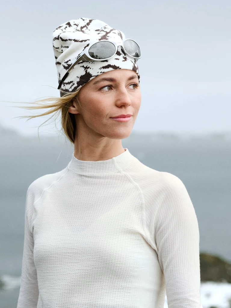 Blonde Frau, die nach rechts schaut, trägt ein weißes Thermoshirt und eine gemusterte Beanie mit Sonnenbrille. Ihr Haar weht im Wind.