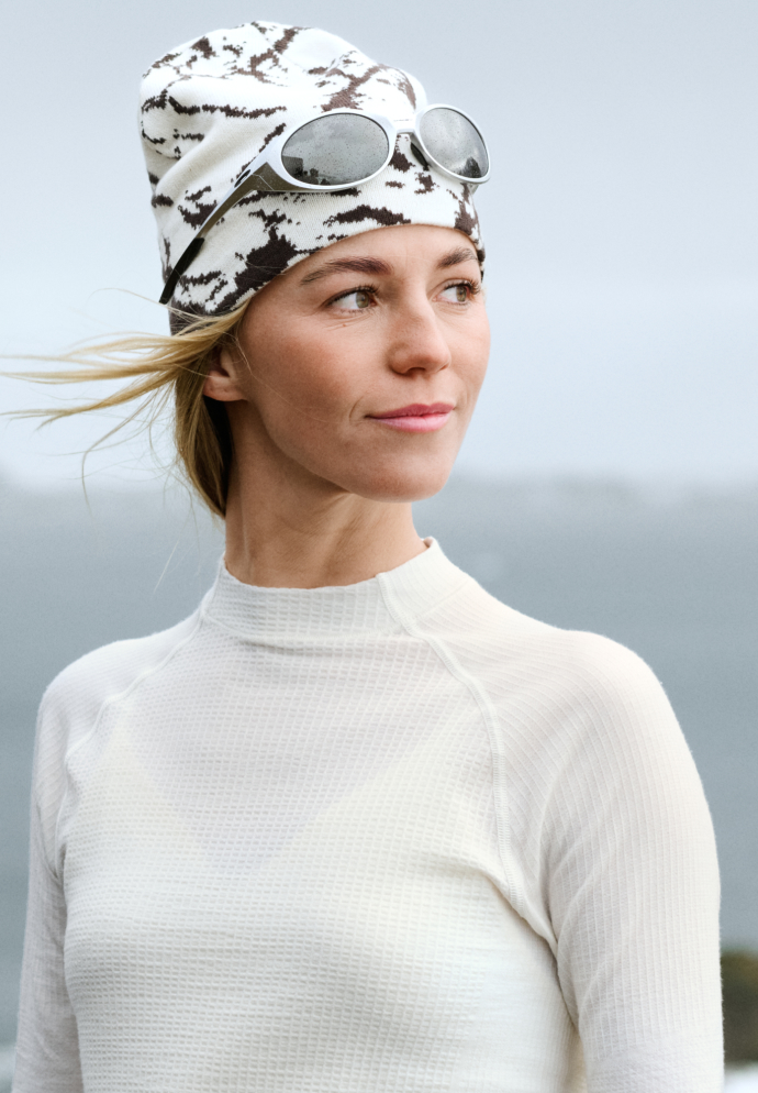 Blonde vrouw met in de wind wapperende haren, gekleed in een witte thermische top en een gemusterde beanie met een zonnebril, die opzij kijkt.