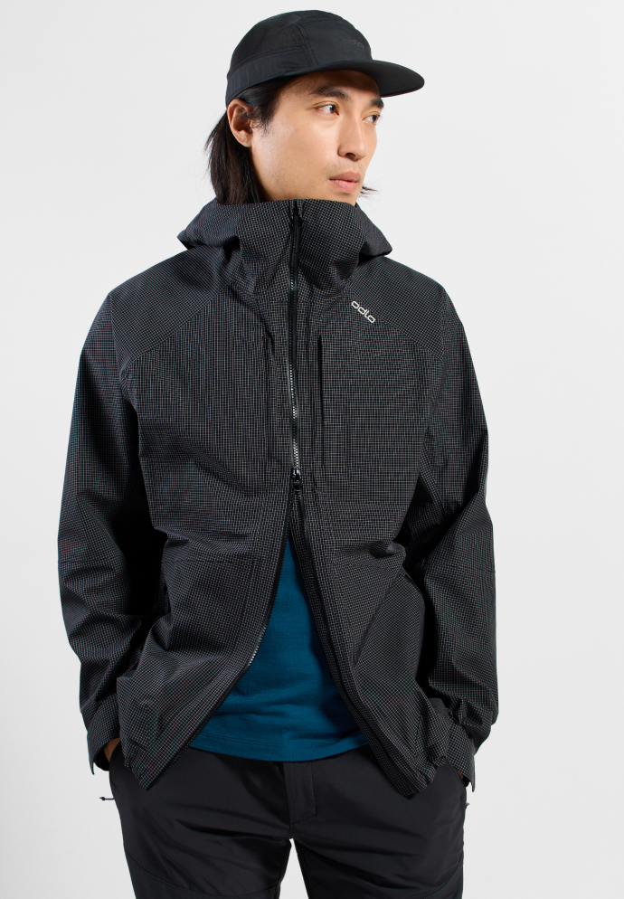 Man wearing a black cap, dark patterned Odlo hooded jacket, blue t-shirt, and black trousers against a white background.