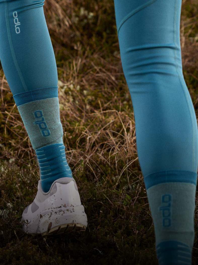 Legs in blue Odlo leggings and socks, wearing muddy white running shoes, walking on grassy outdoor terrain.