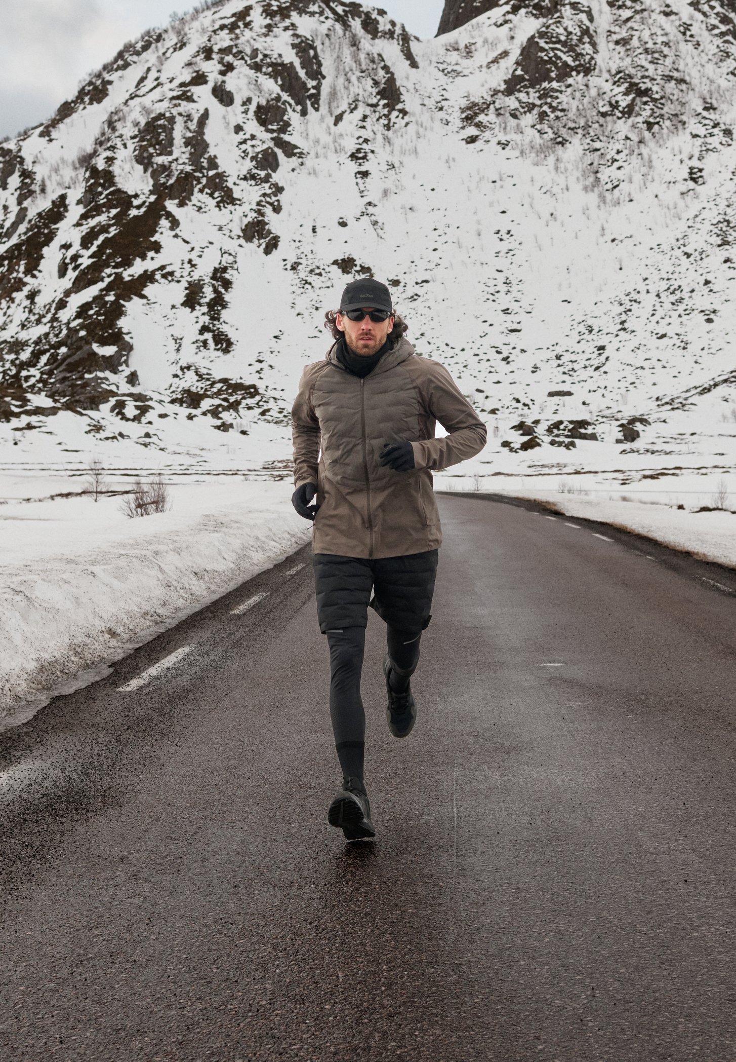 Un uomo con una giacca marrone e leggings neri corre su una strada bagnata con montagne innevate sullo sfondo.