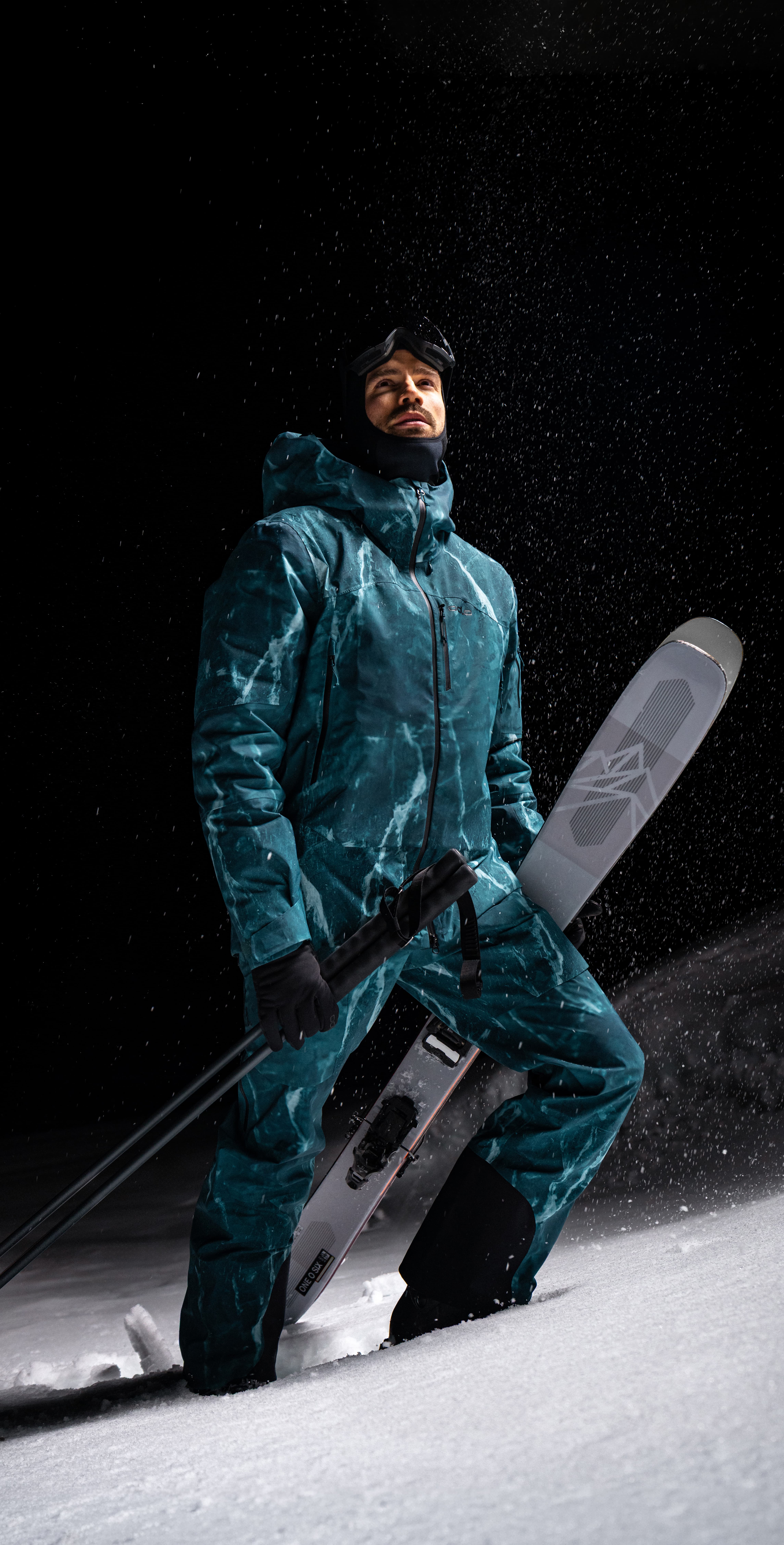 Man in a teal ski suit holding skis and poles, standing on a snowy slope at night looking up at falling snow.