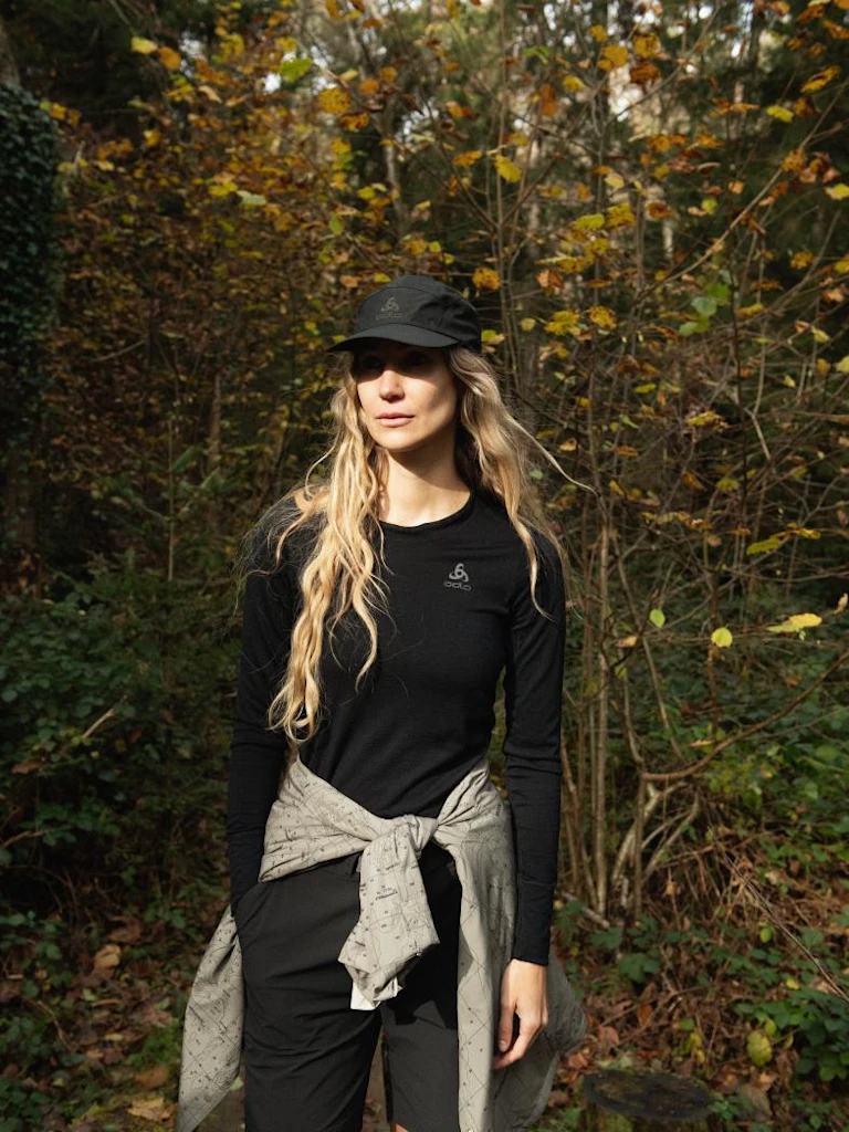 Blonde woman in a black Odlo cap and long-sleeve base layer stands in an autumn forest with a jacket tied around her waist.