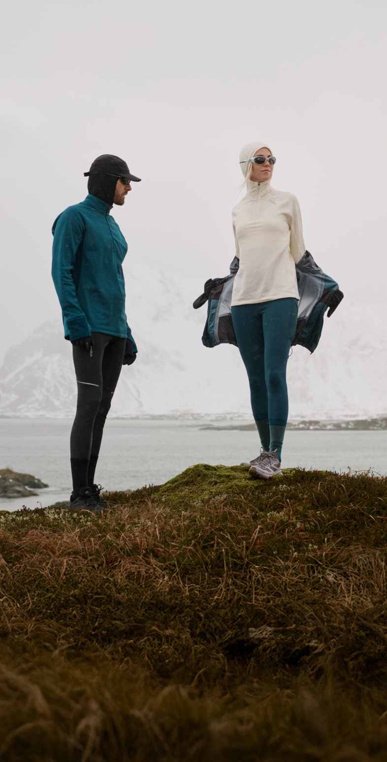 Two people in winter layers stand on a grassy hill by a lake and snowy mountains. The woman is taking off her dark jacket showing the mid layer under.