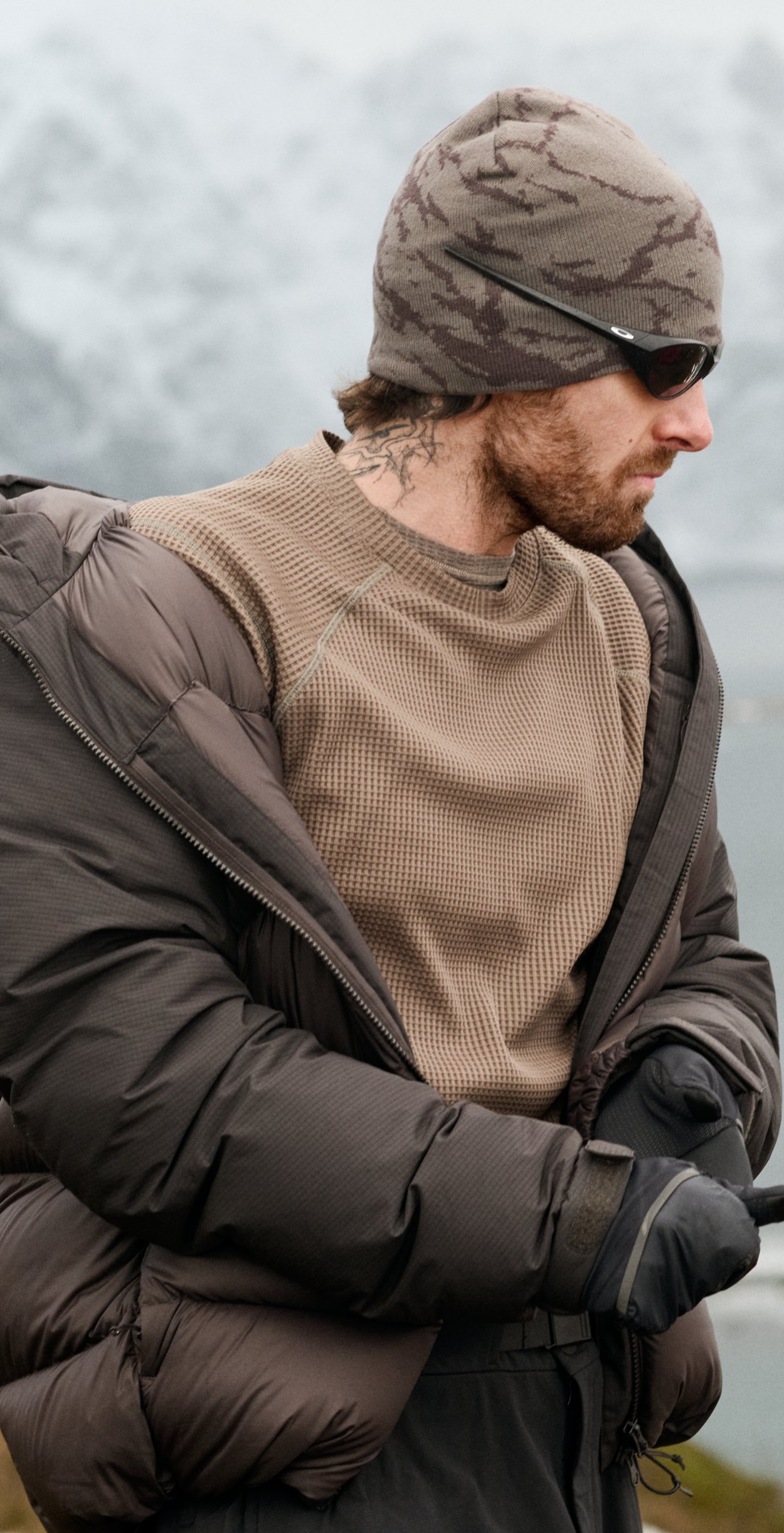 Bärtiger Mann in brauner Jacke, Camouflage-Beanie und Sonnenbrille richtet seine Handschuhe vor schneebedeckten Bergen und Wasser.
