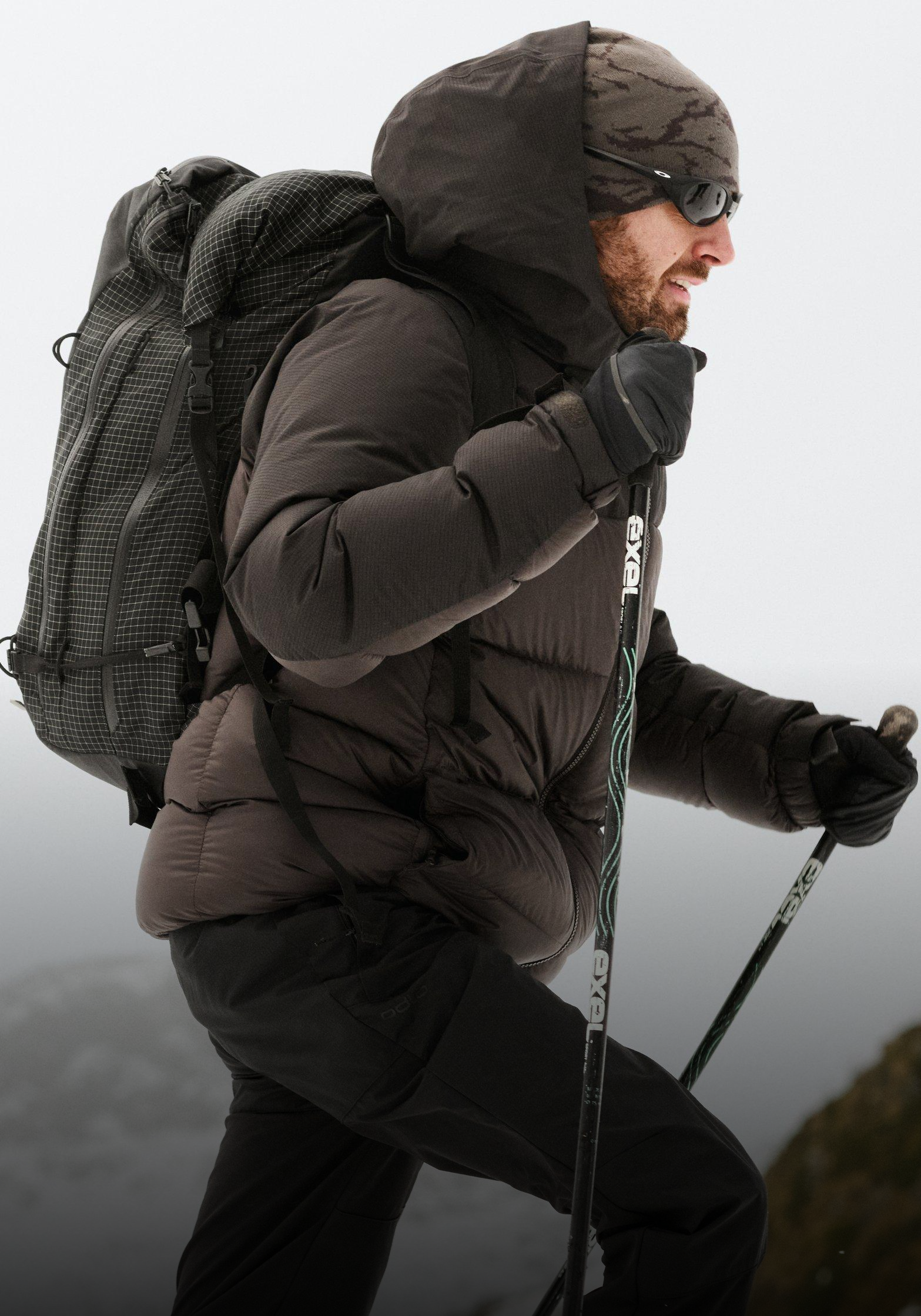 Profiel van een man met baard in een donker jack met capuchon, camogekleurde beanie, zonnebril en een zwarte rugzak met grid-patroon tegen een grijze lucht.