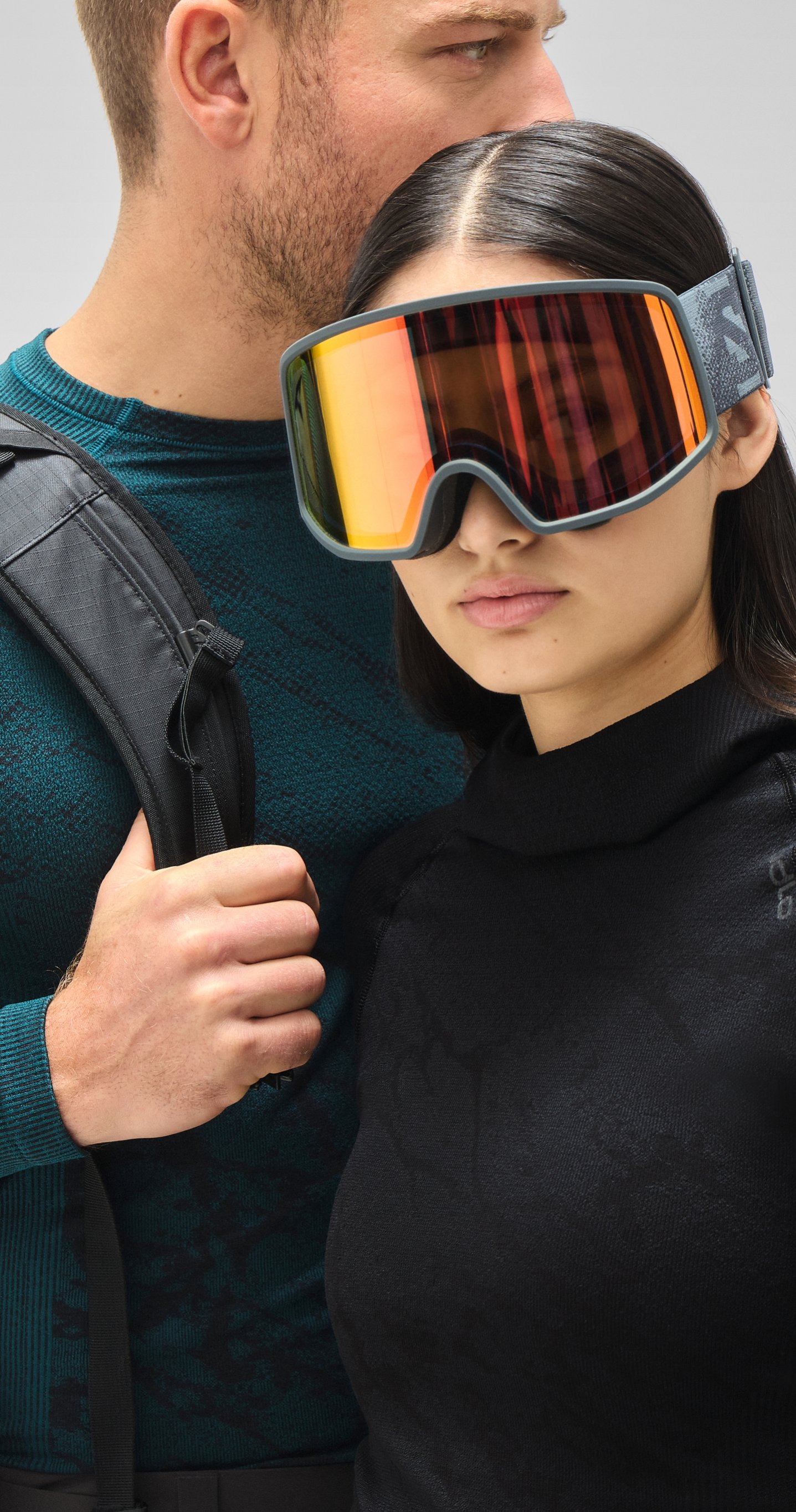 Woman in reflective orange ski goggles and black top stands by a man in a teal patterned shirt holding a black backpack.