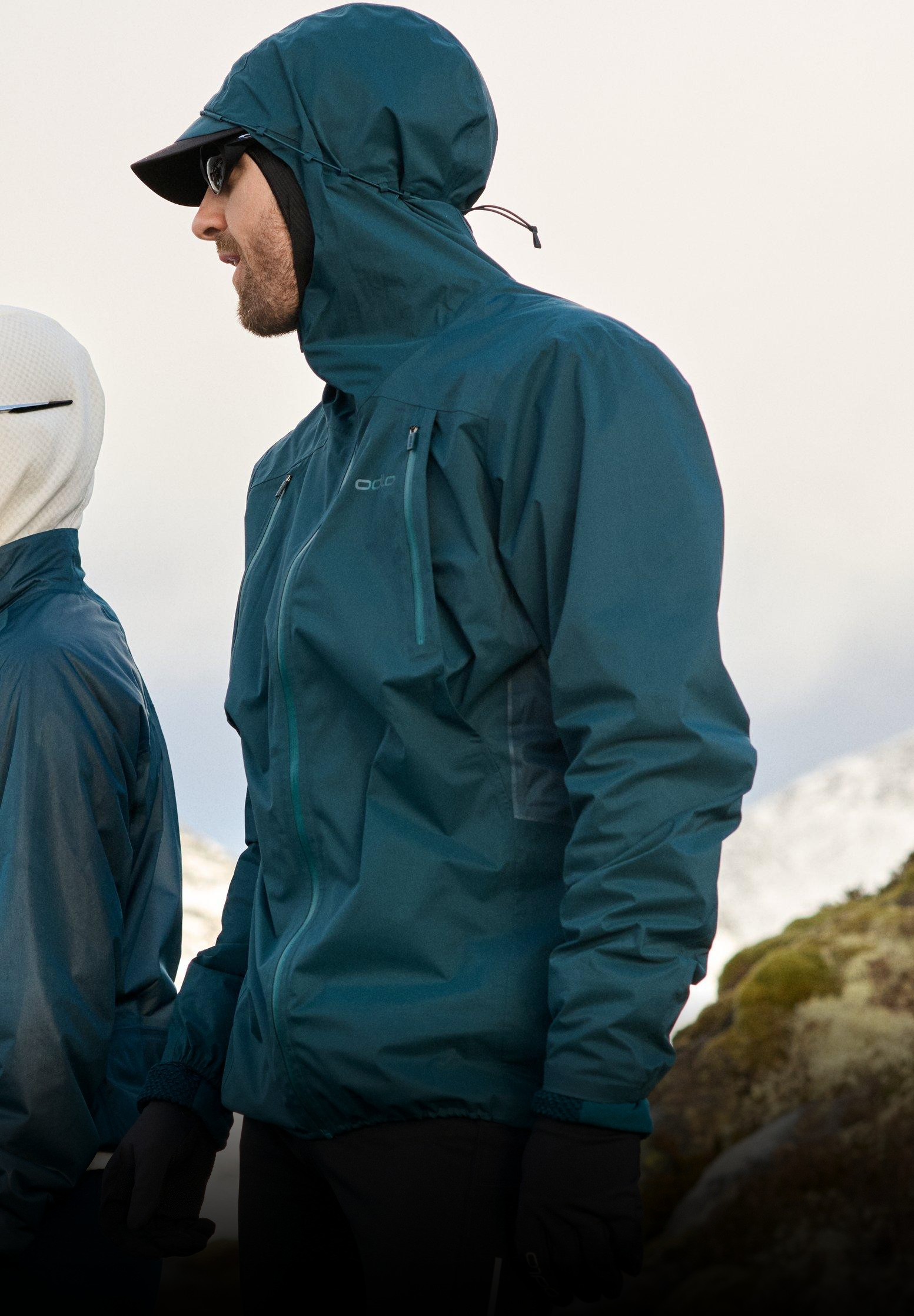 Mann in einer dunkeltürkisen Kapuzenjacke, mit Cap, Sonnenbrille und Handschuhen, steht im Freien vor einer schneebedeckten Bergkulisse.