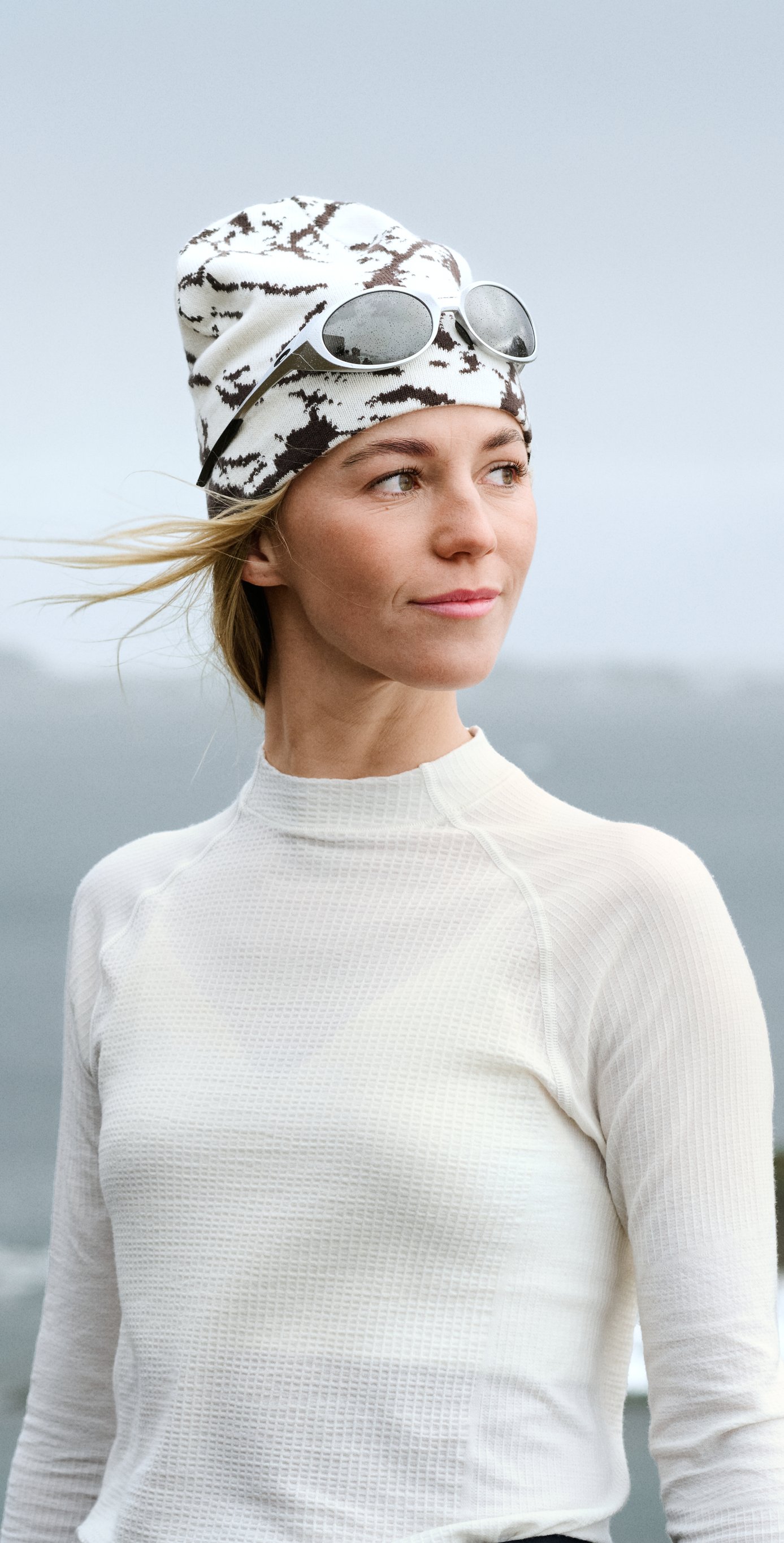 Woman in white thermal top and patterned beanie with sunglasses on head, looking right with wind blowing her blonde hair.