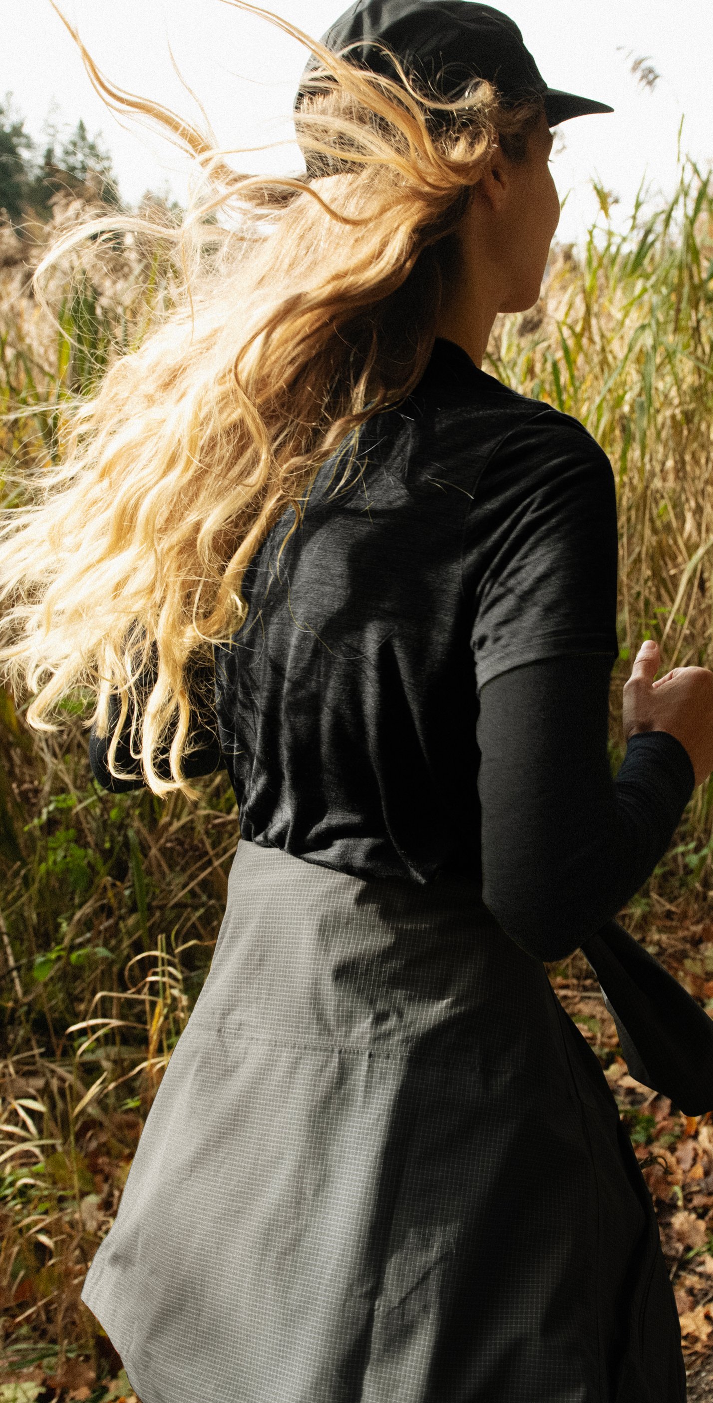 Blonde Frau in Schwarz beim Running auf einem Pfad durch hohes, trockenes Gras, ihr langes Haar weht im Wind.