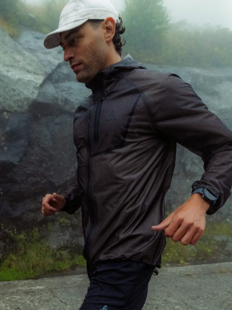 A man running in the rain wearing a dark jacket, blue shorts, and a white cap against a foggy, rocky background.