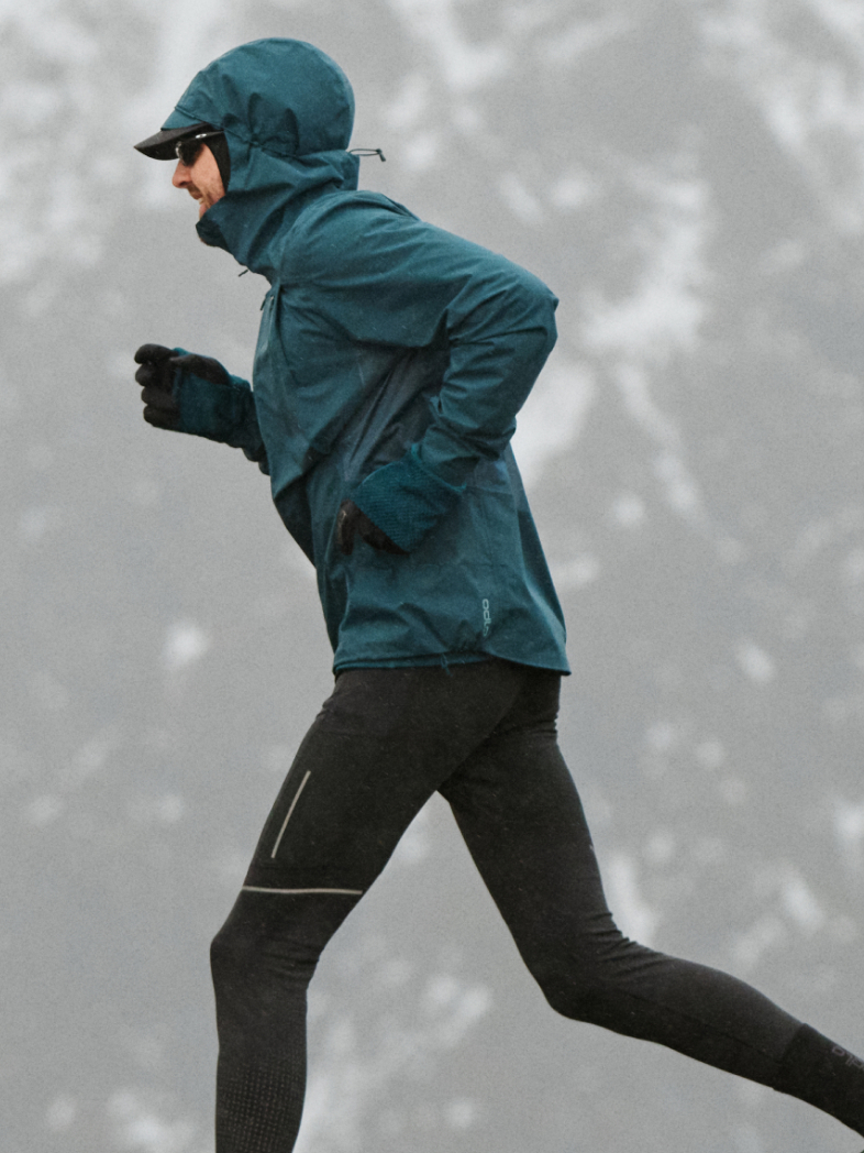 A person running in a snowy landscape wearing a teal hooded jacket, black running tights, gloves, and sunglasses.
