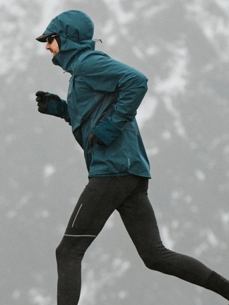 A person running in a snowy landscape wearing a teal hooded jacket, black running tights, gloves, and sunglasses.