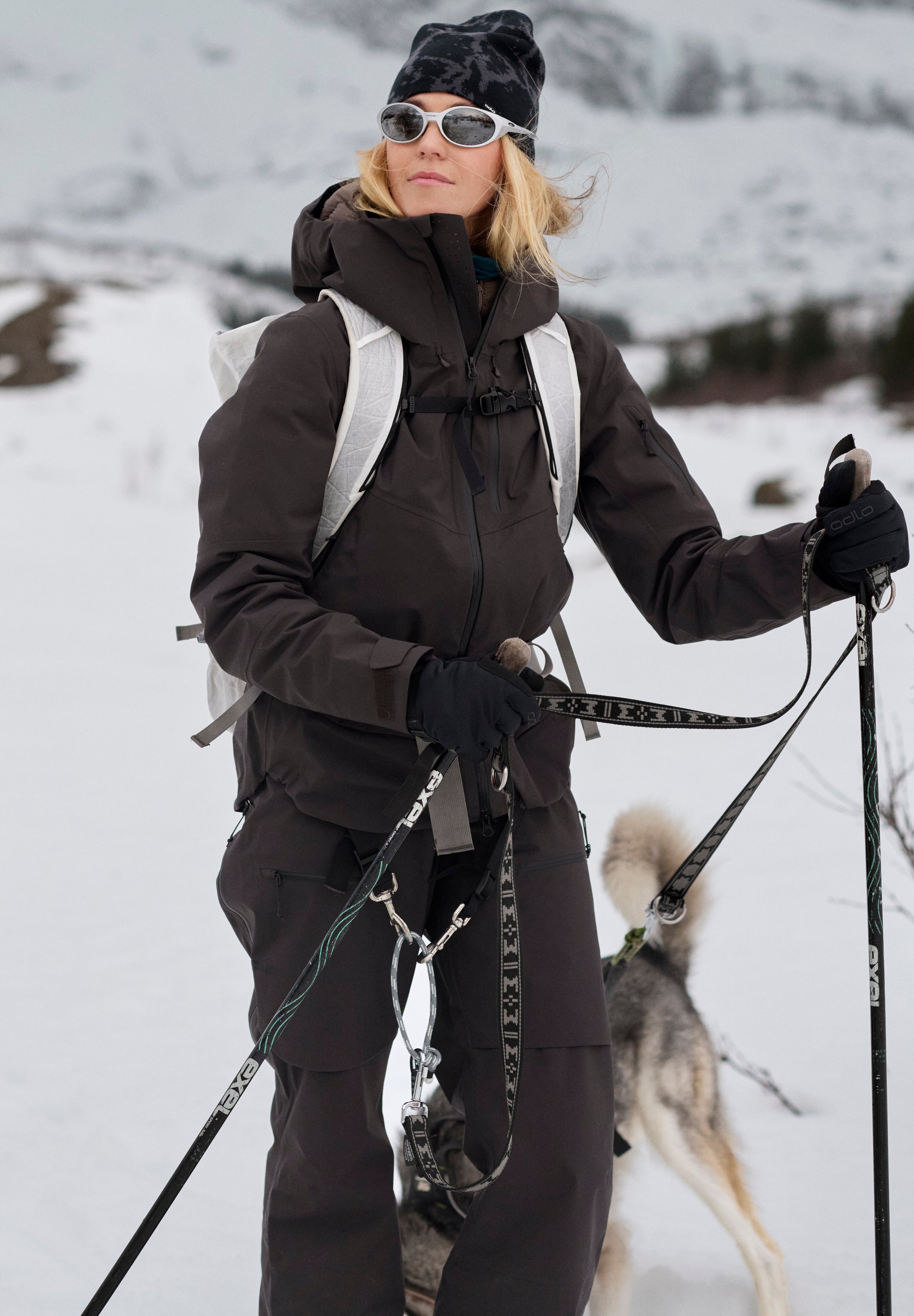 Blonde Frau in dunkler Winterausrüstung und Sonnenbrille steht im Schnee und hält Skistöcke, mit einem Husky an der Leine hinter ihr.
