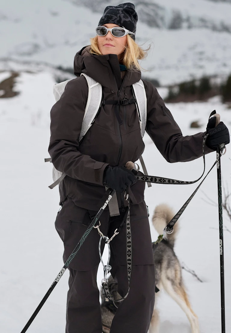Blonde woman in dark winter gear and sunglasses stands in snow holding ski poles, with a husky dog on a leash behind her.
