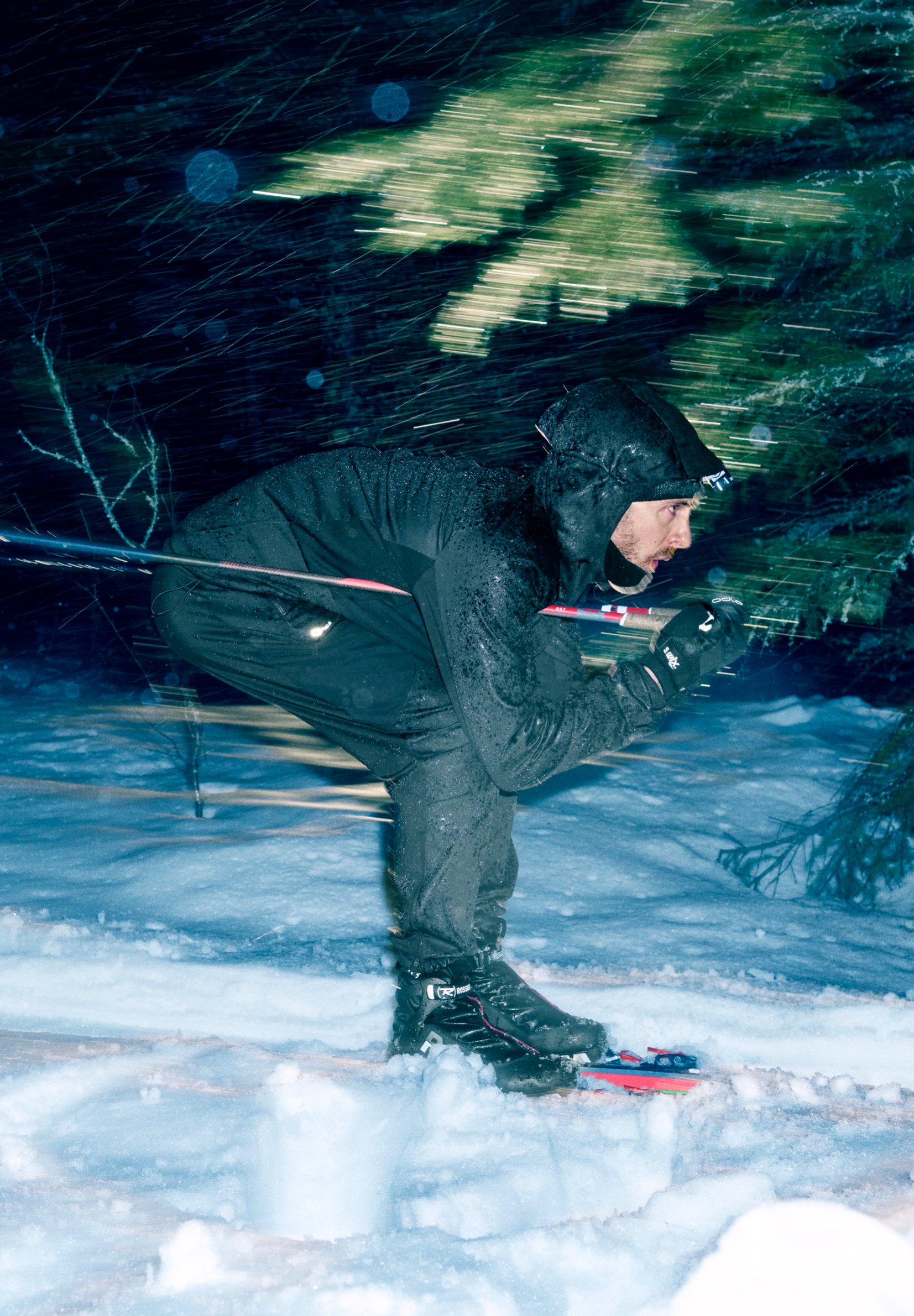 Un homme en tenue noire file à toute allure sur une piste de ski de fond enneigée en pleine nuit, penché en avant, bravant une violente tempête de neige.