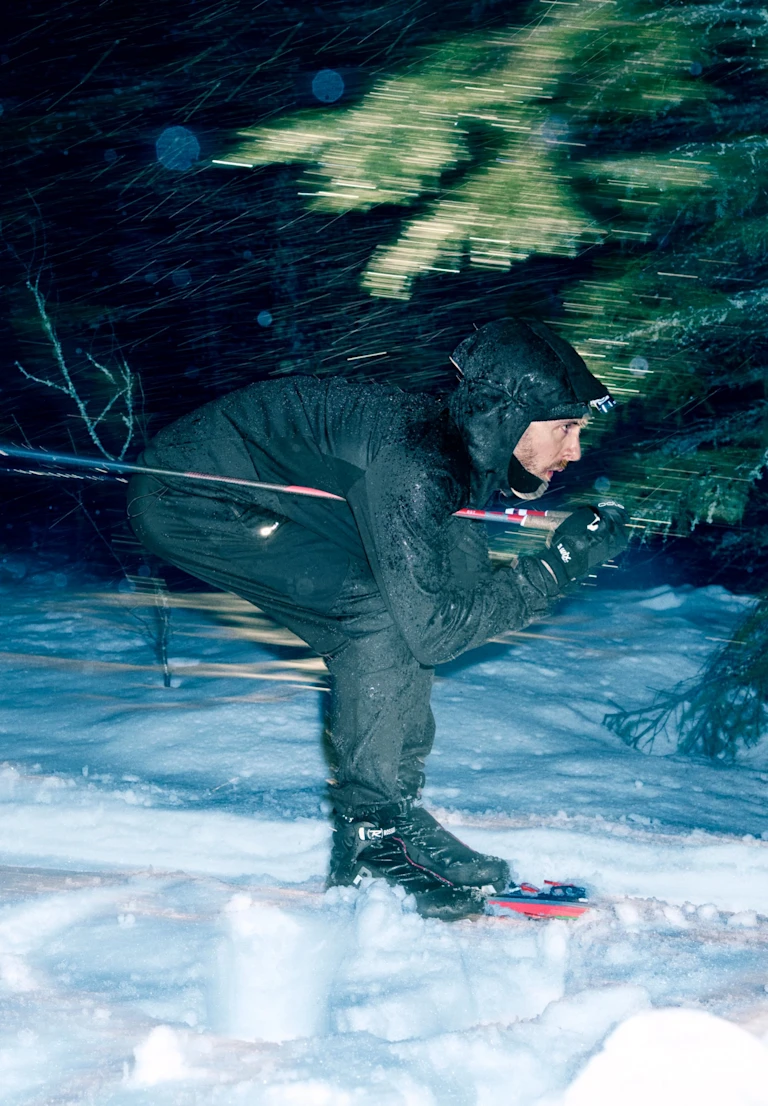 Un homme en tenue noire file à toute allure sur une piste de ski de fond enneigée en pleine nuit, penché en avant, bravant une violente tempête de neige.