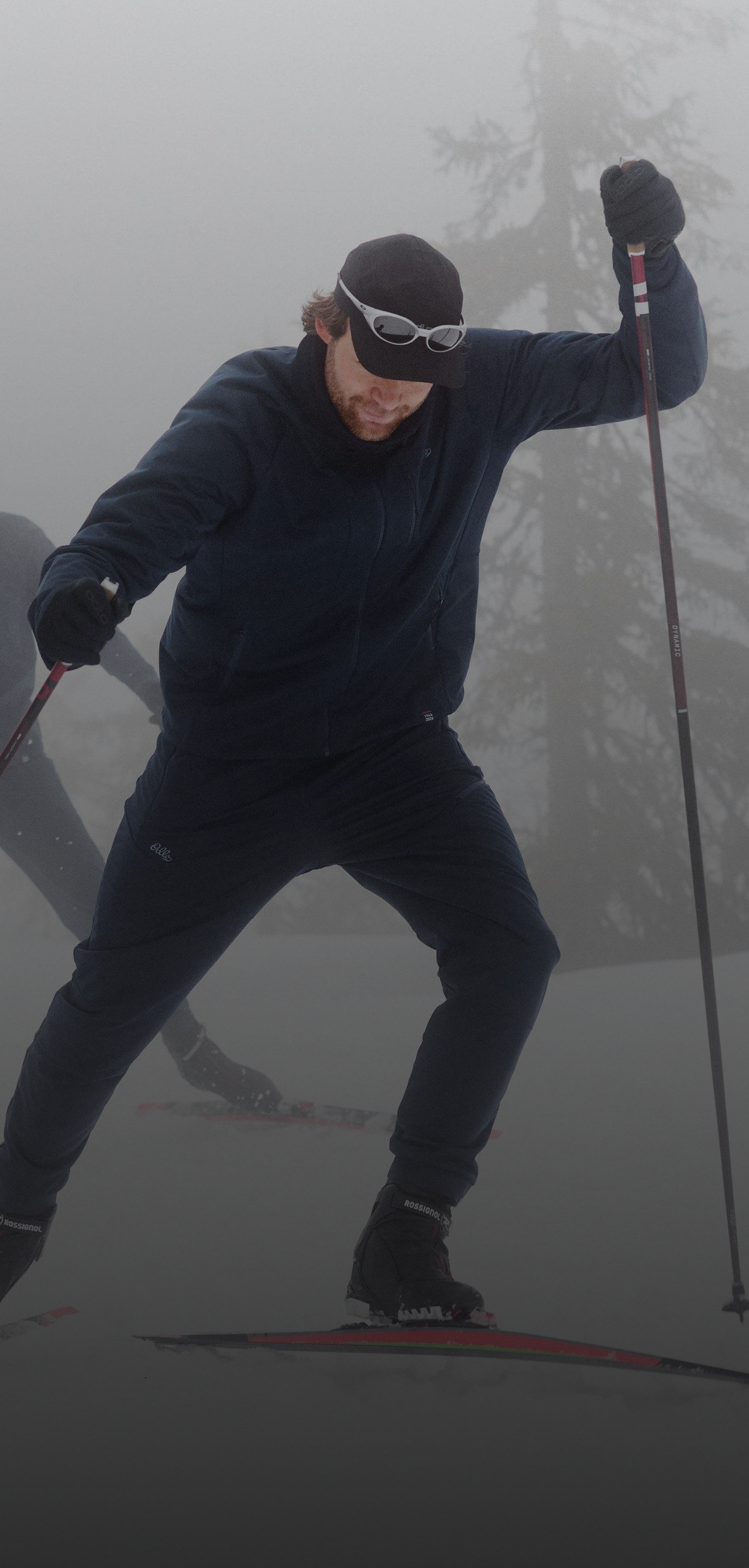 Zwei Langläufer gleiten durch den dichten Nebel eines verschneiten Waldes, gekleidet in dunkle Langlauf Sportbekleidung und Sonnenbrillen.