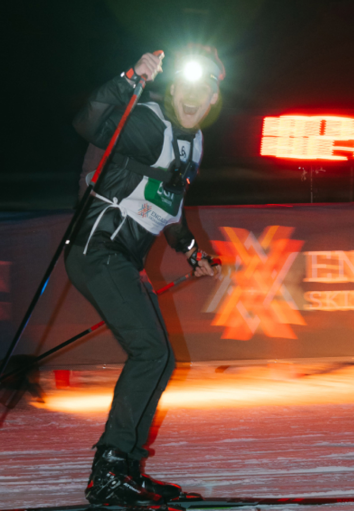 Smiling cross-country skier racing at night with a bright headlamp, dark clothing, and a white race bib on snow.