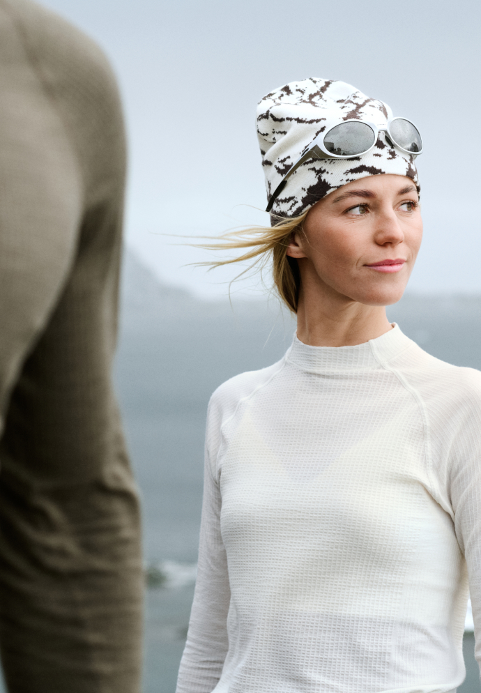 Frau in weißem Thermo-Shirt und gemusterter Beanie mit Sonnenbrille auf dem Kopf, blickt nach rechts, während der Wind durch ihr blondes Haar weht.