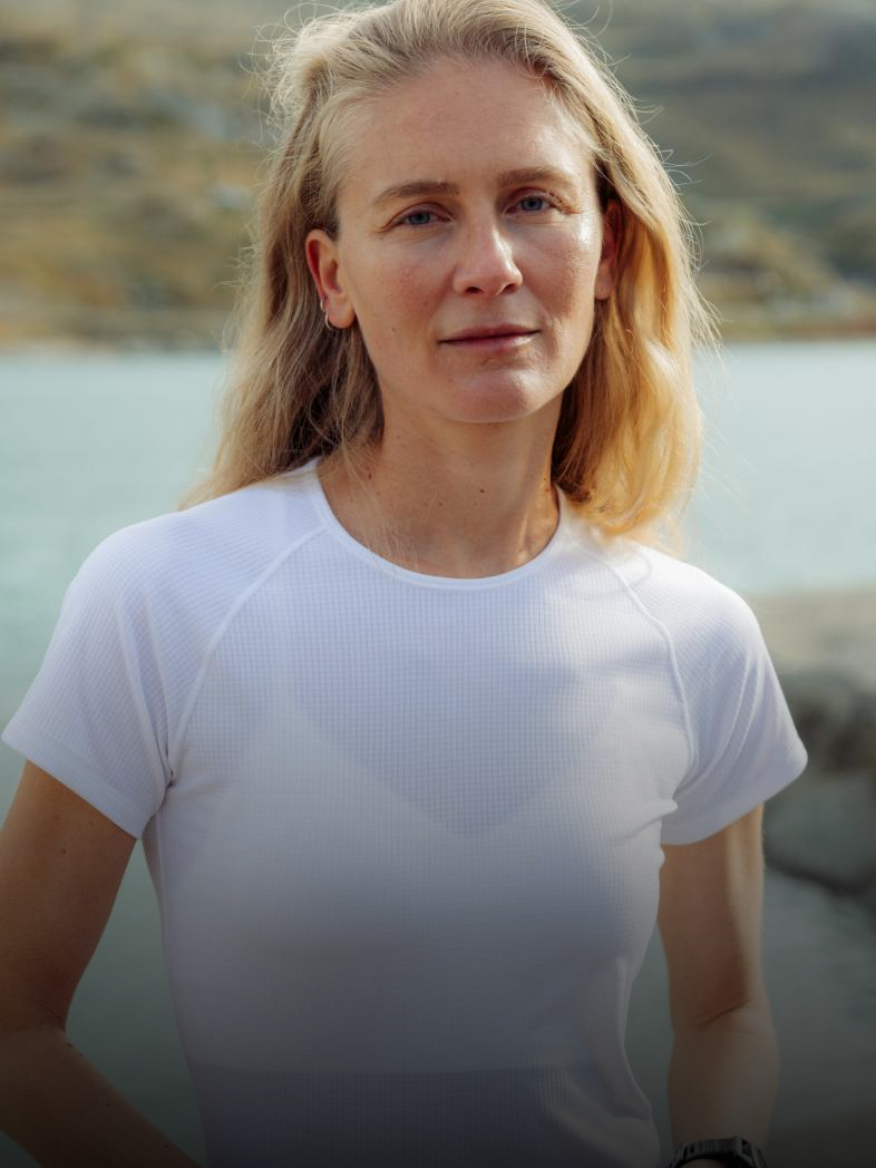 Donna bionda con un base layer bianco leggero che guarda verso la fotocamera, con un lago e colline rocciose sfocati sullo sfondo.