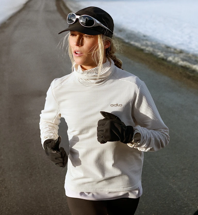 Donna bionda che corre su una strada asfaltata fiancheggiata dalla neve, con indosso un top Odlo bianco, un berretto nero, occhiali da sole e guanti neri.