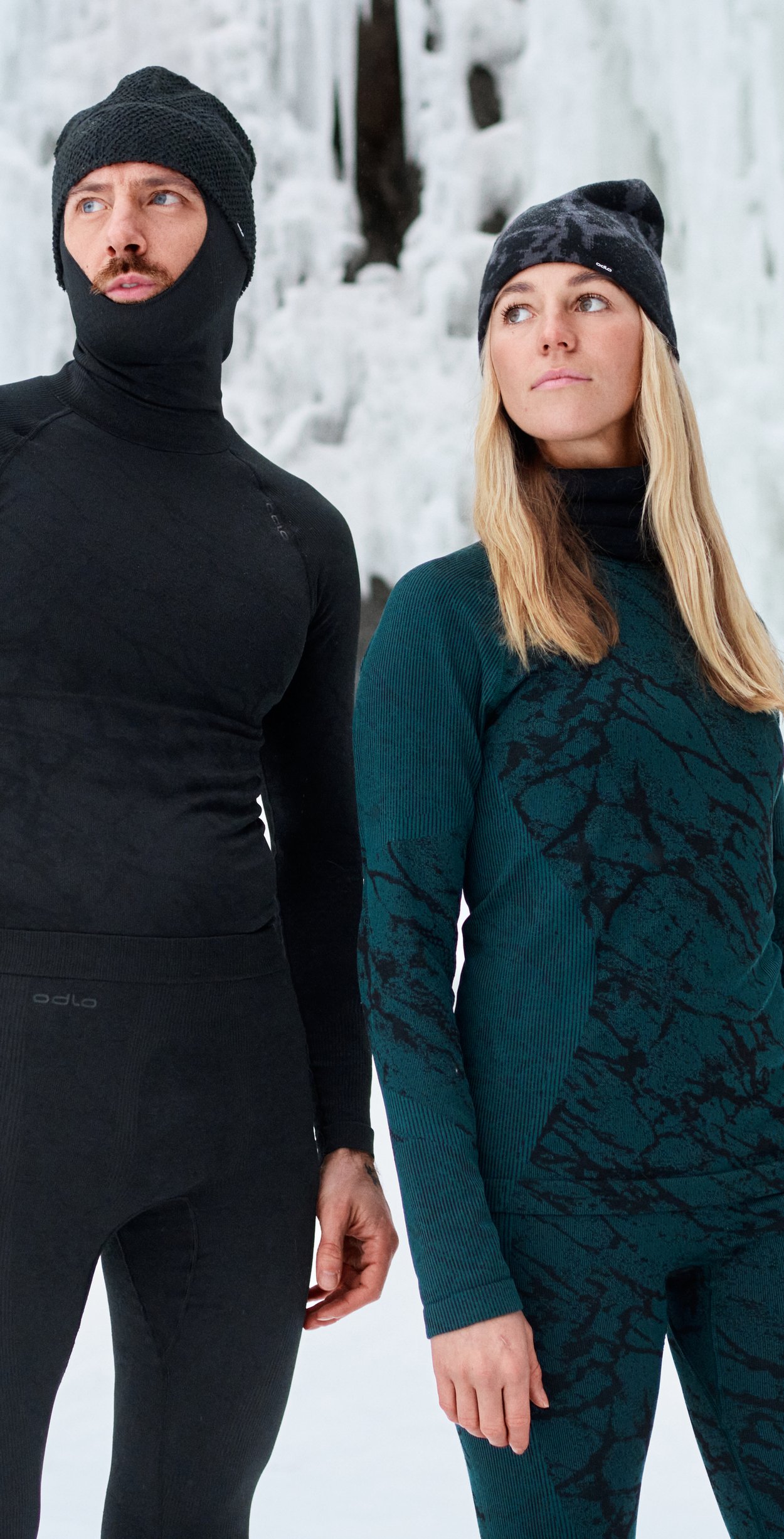Man in zwarte thermokleding met capuchon en vrouw in groen gepatroneerde thermokleding staan in de sneeuw voor een bevroren waterval.