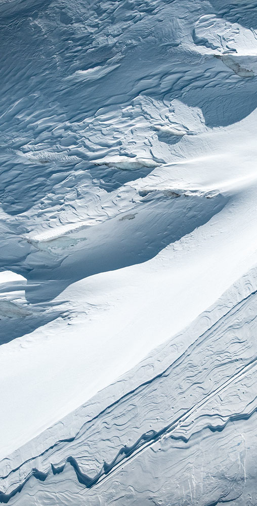 Aerial view of two skiers ascending a steep, textured white snow ridge, casting long dark shadows on the mountain.