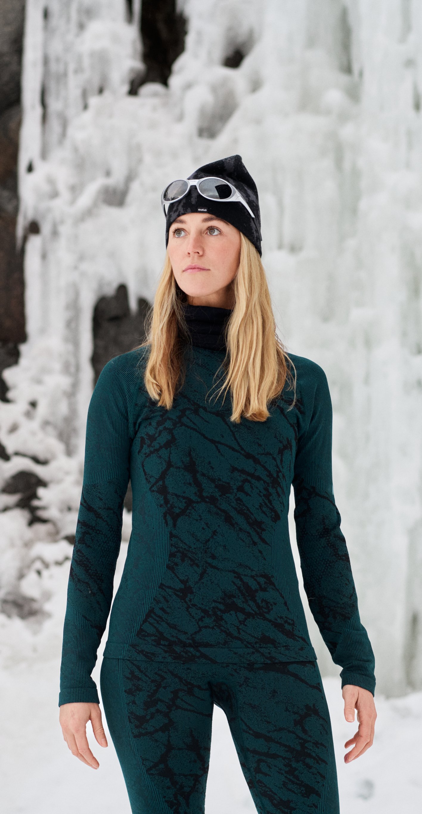 Woman in teal thermal wear and beanie with goggles stands in a snowy landscape before towering icicles and dark rock.