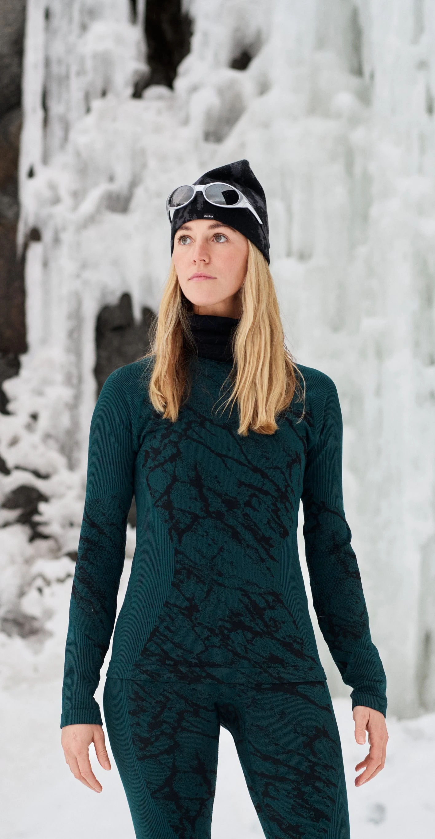 Woman in teal thermal wear and beanie with goggles stands in a snowy landscape before towering icicles and dark rock.
