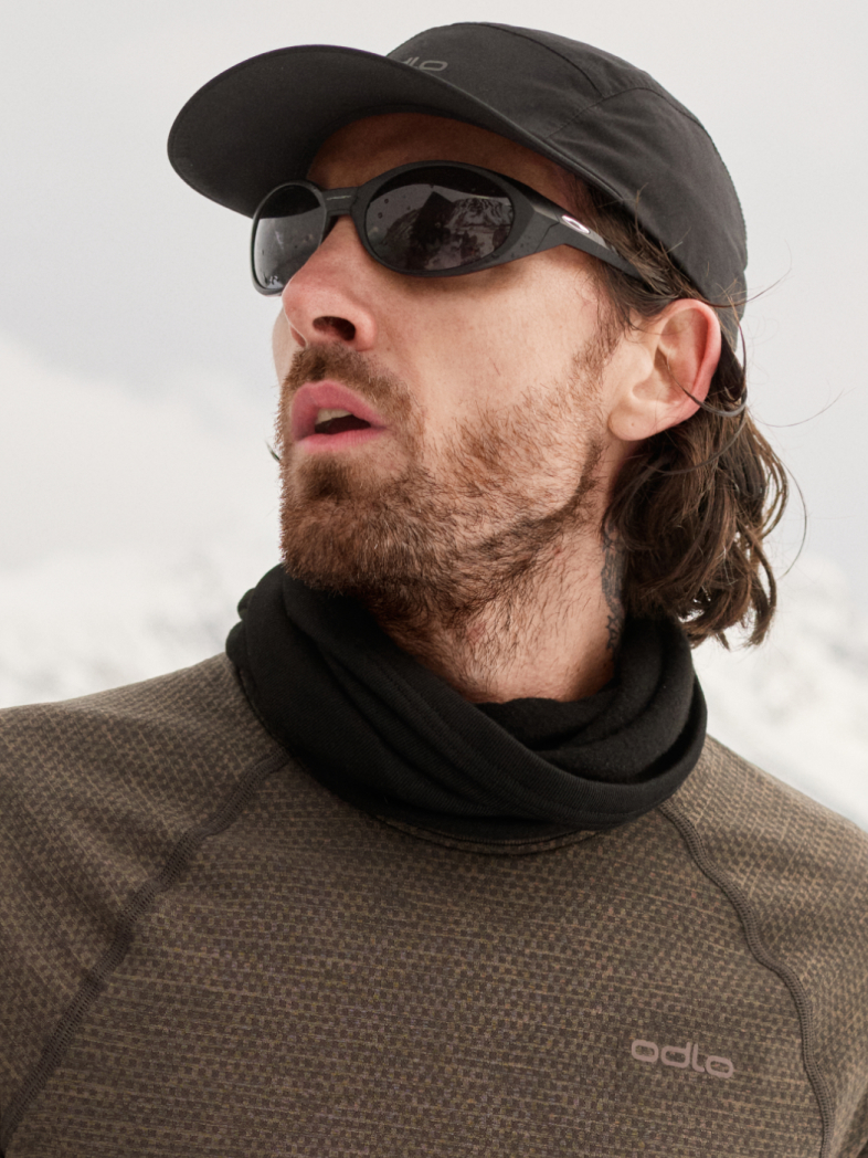 Man in black cap and sunglasses wearing a neck gaiter and brown thermal top, looking left against a snowy mountain backdrop.
