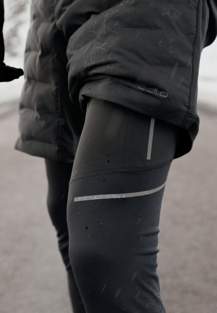 Close-up of a person's legs in black quilted shorts and black tights with reflective stripes, covered in water droplets.