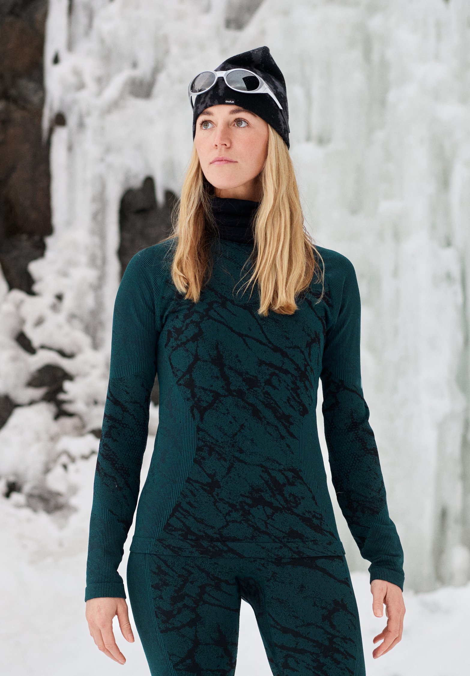 Woman in teal patterned activewear and black beanie with goggles standing before a frozen waterfall backdrop.