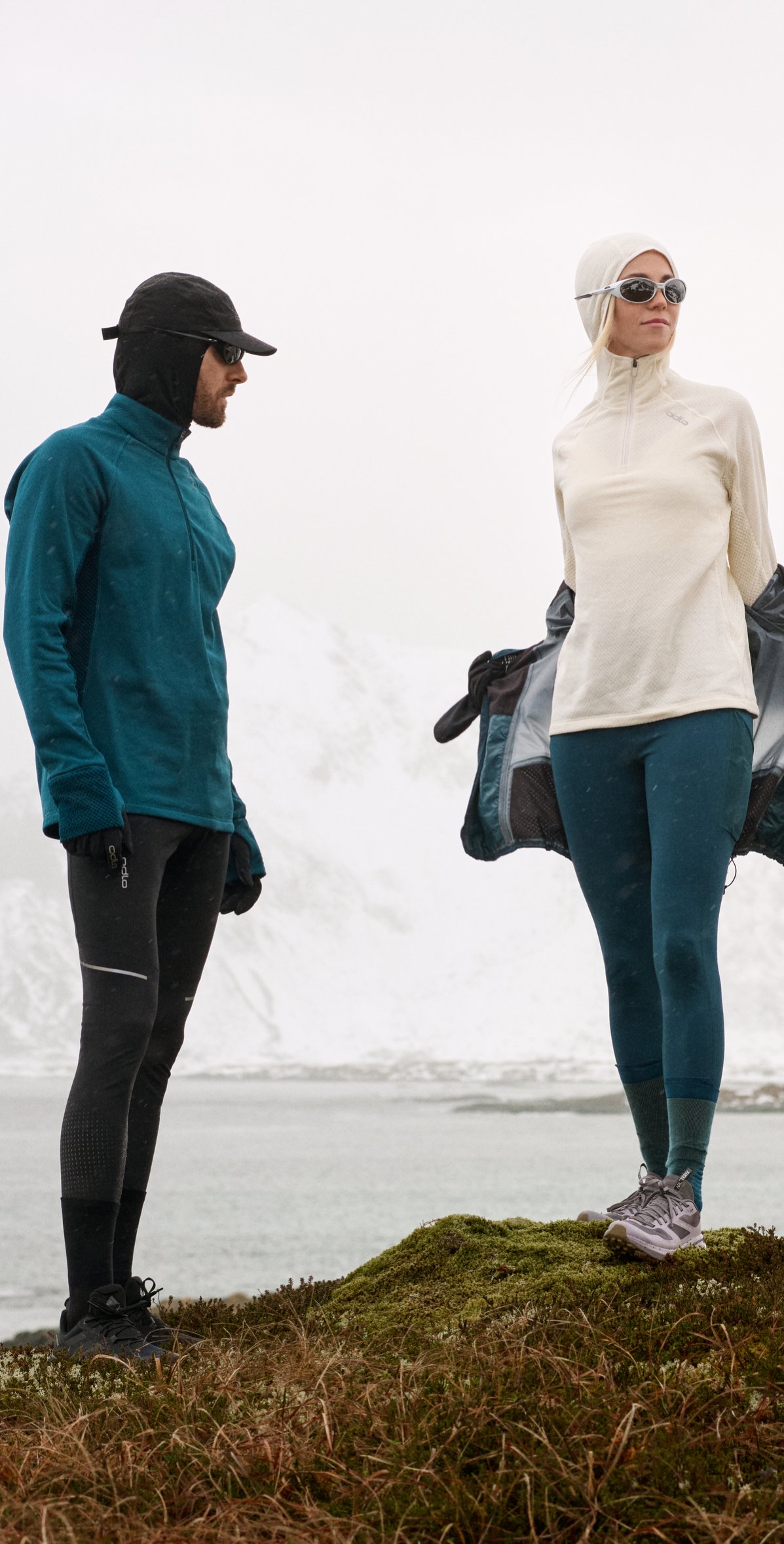 Twee hardlopers in winterkleding staan op een met mos begroeide heuvel bij een besneeuwde fjord; de een draagt een petrolkleurige jas en pet, de ander een crèmekleurige top en een zonnebril.