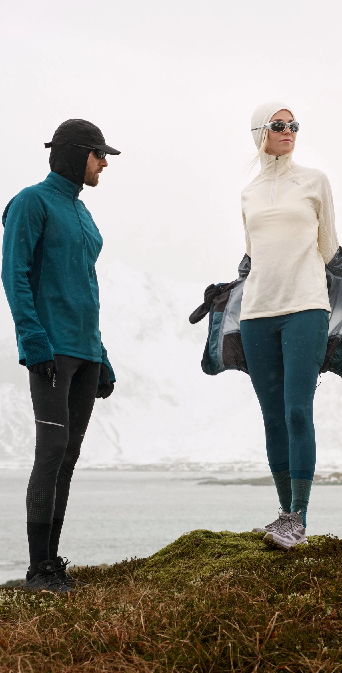 Twee hardlopers in winterkleding staan op een met mos begroeide heuvel bij een besneeuwde fjord; de een draagt een petrolkleurige jas en pet, de ander een crèmekleurige top en een zonnebril.