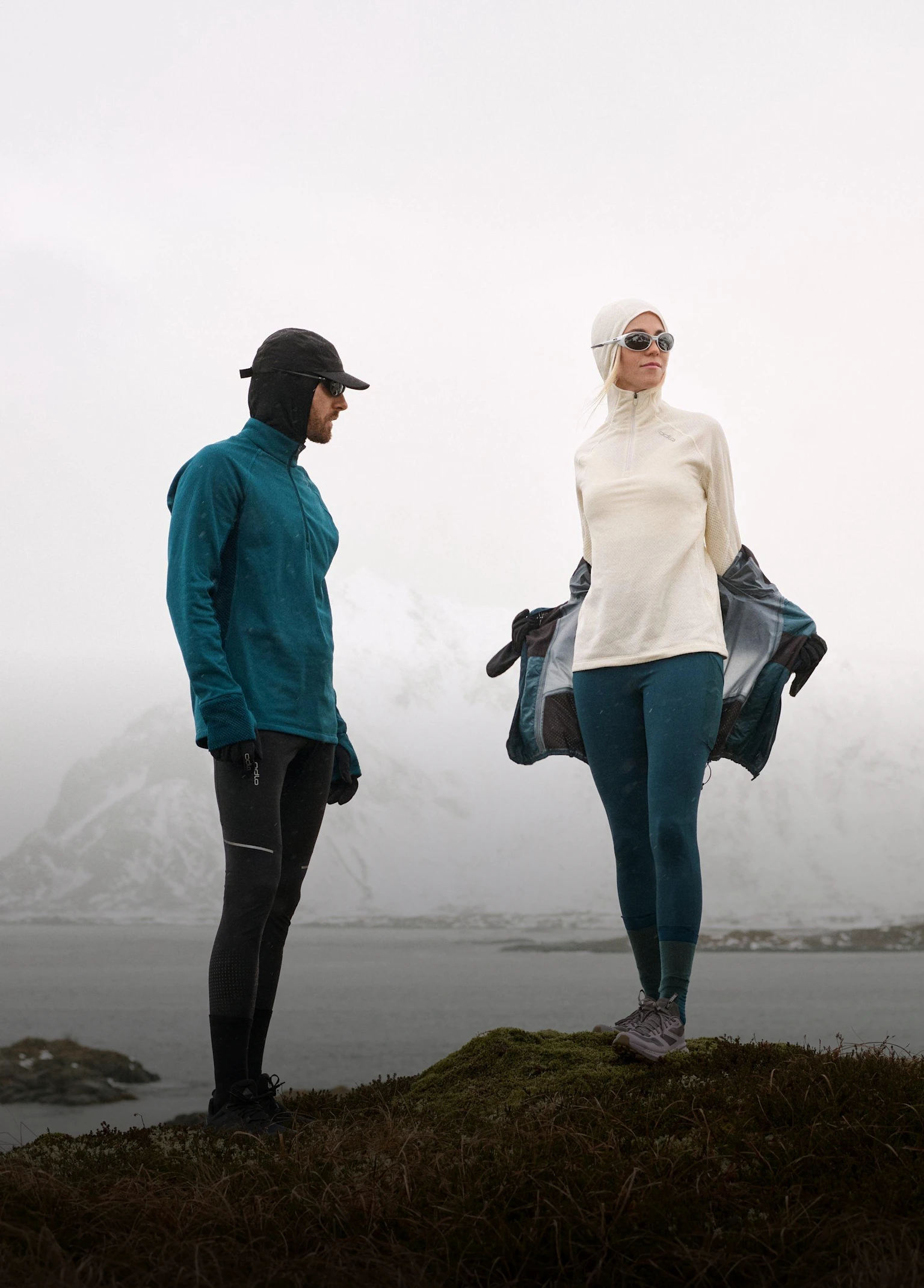 Two people in winter athletic gear stand in a snowy landscape. The man wears teal; the woman is removing her dark jacket.