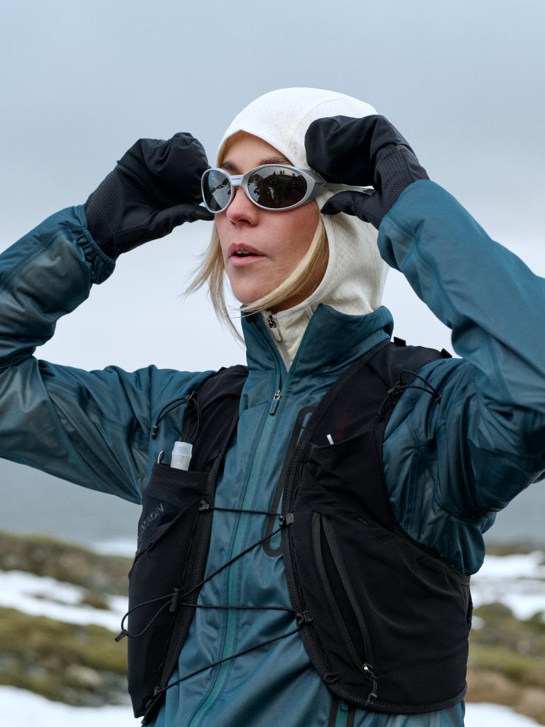 Vrouw in een blauwgroene jas, zwart hardloopvest en witte capuchon past haar zonnebril aan met behandschoende handen in een besneeuwd landschap.
