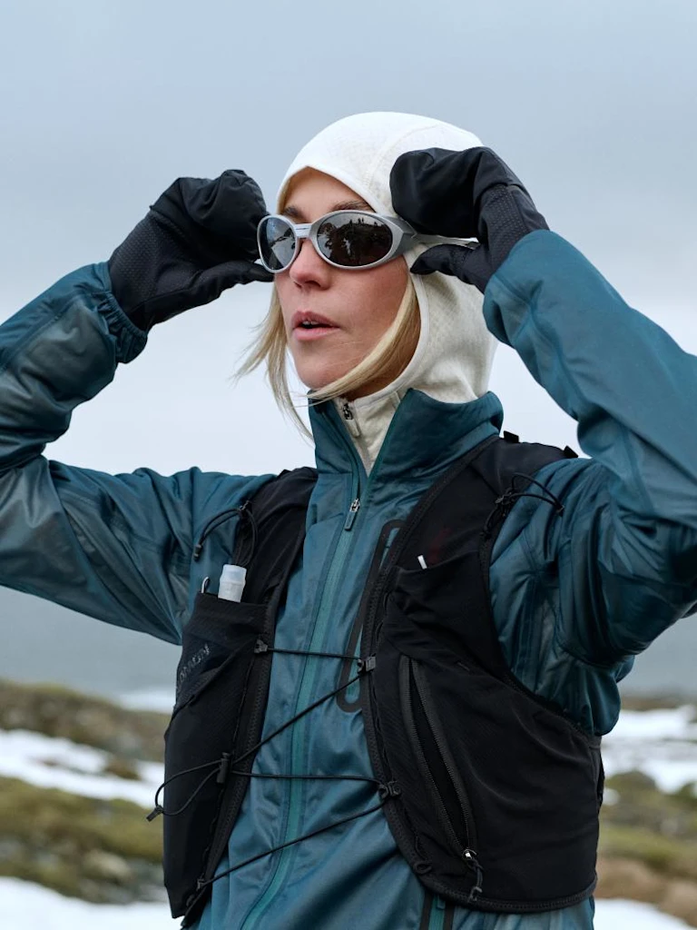 Vrouw in een blauwgroene jas, zwart hardloopvest en witte capuchon past haar zonnebril aan met behandschoende handen in een besneeuwd landschap.
