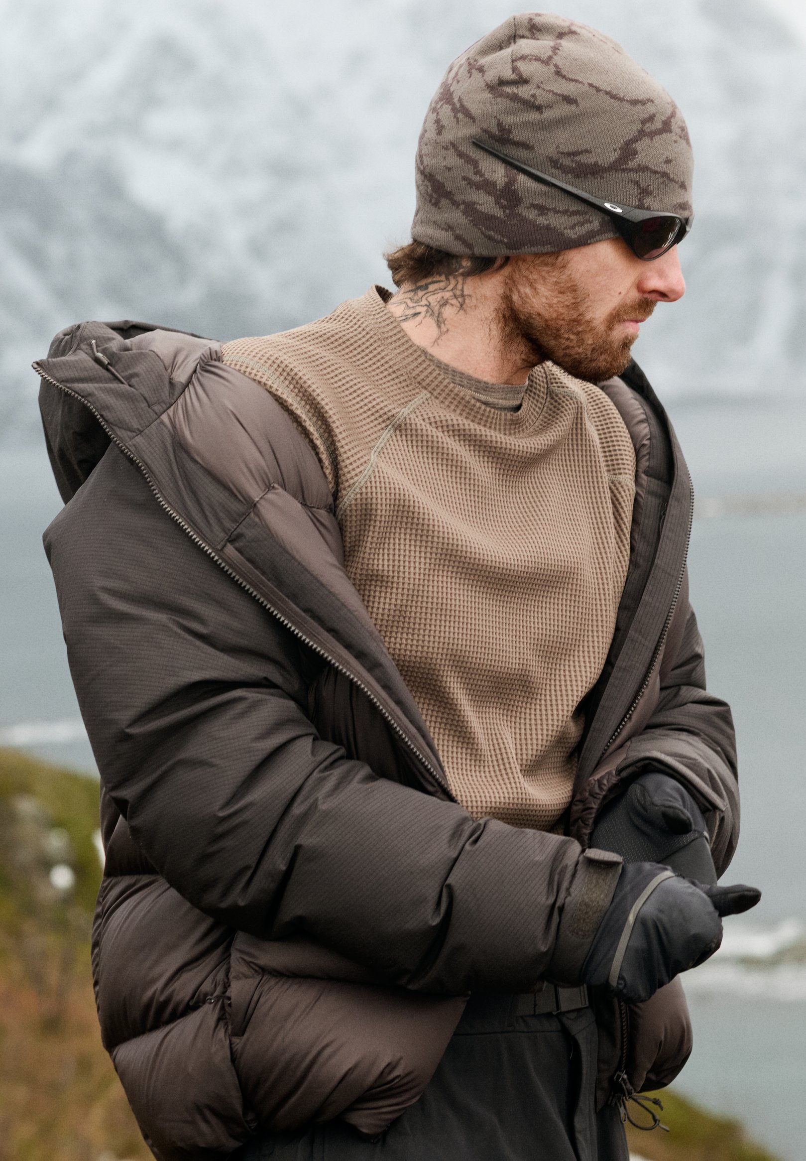 Bearded man in a brown puffer jacket, camo beanie and sunglasses adjusts his gloves in a snowy, mountainous landscape.