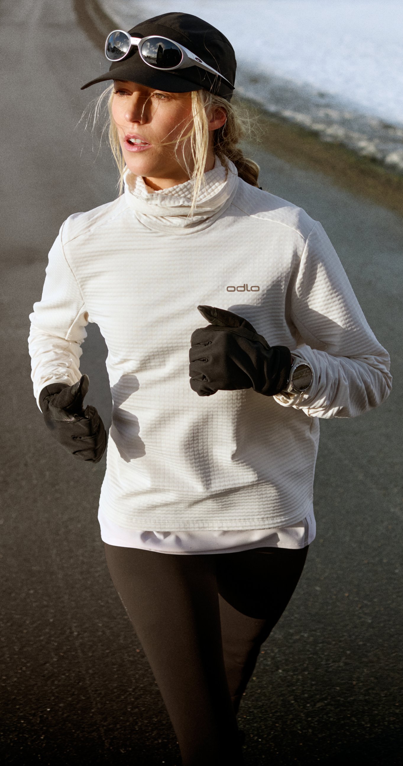 Eine Frau beim Running auf einer asphaltierten Straße, umgeben von Schnee, in einem weißen Odlo Top, mit schwarzer Cap, Sonnenbrille und schwarzen Handschuhen.
