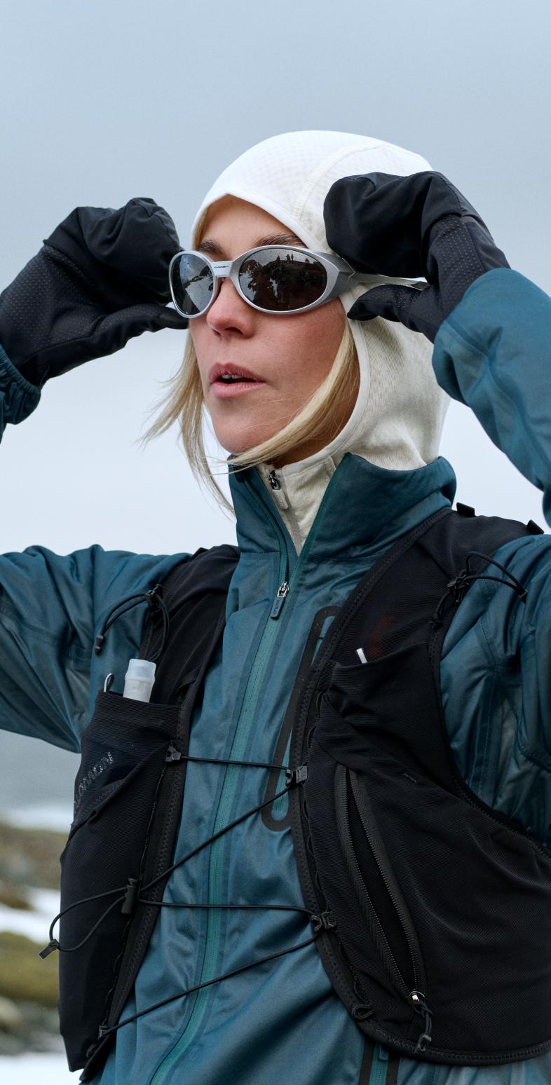 Une femme en veste bleue, capuche blanche et gilet noir ajuste ses lunettes de soleil avec des mains gantées, dans un paysage enneigé et rocailleux.