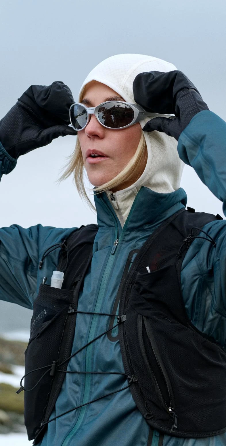 Une femme en veste bleue, capuche blanche et gilet noir ajuste ses lunettes de soleil avec des mains gantées, dans un paysage enneigé et rocailleux.