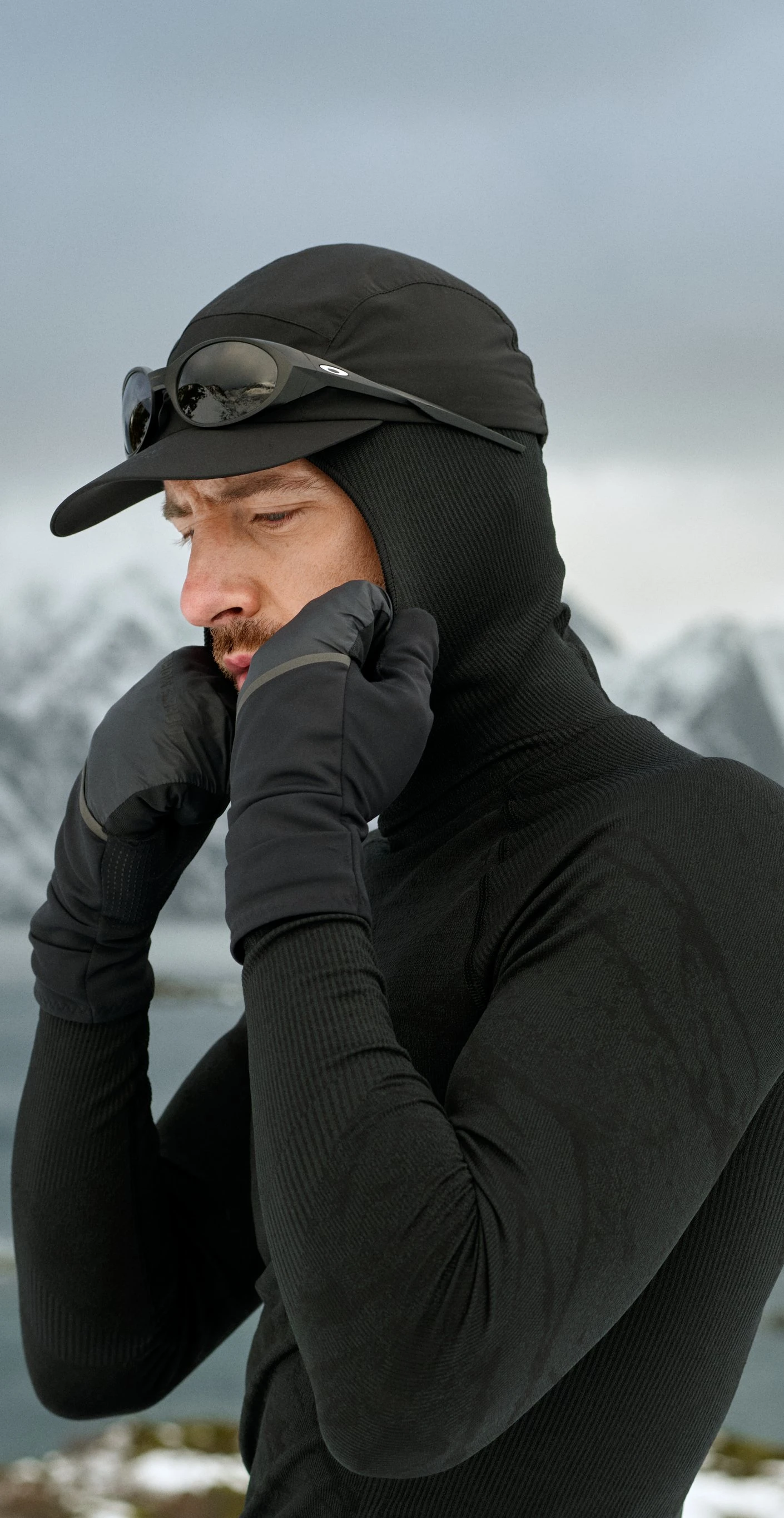 Mann in schwarzer Thermobekleidung, mit Handschuhen und einer Mütze mit Sonnenbrille, richtet seine Kapuze vor einer verschneiten Berglandschaft.