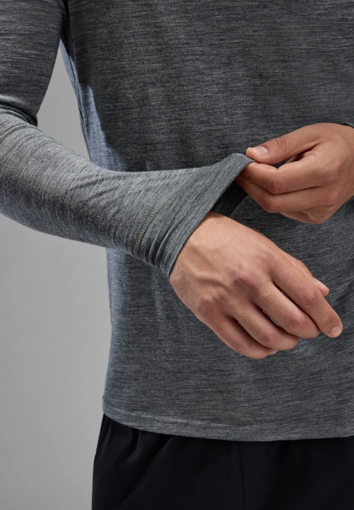 Nahaufnahme eines Mannes in einem grauen langärmligen Merino Base Layer Oberteil, der am dehnbaren Ärmel zieht, vor einem einfarbigen Hintergrund.