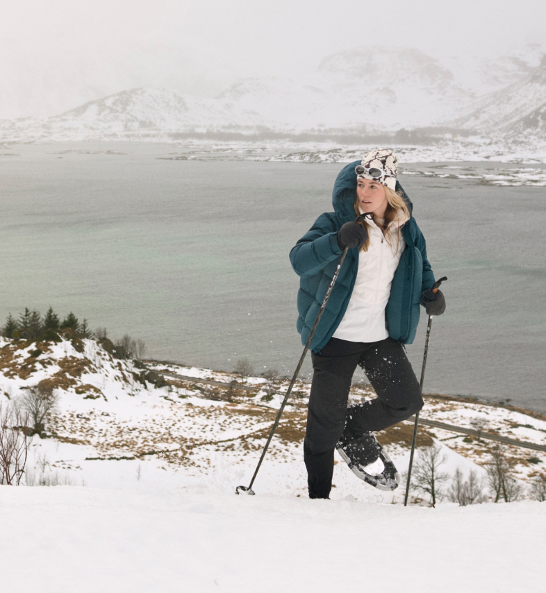 Femme en doudoune sarcelle faisant de la raquette en montée avec des bâtons, surplombant un fjord calme et des montagnes enneigées.