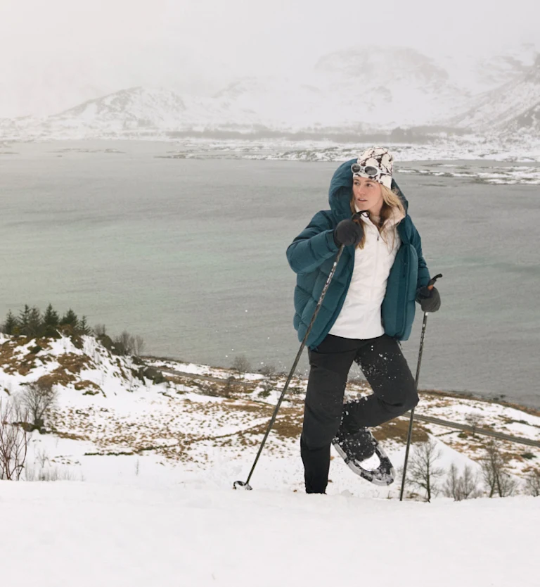 Femme en doudoune sarcelle faisant de la raquette en montée avec des bâtons, surplombant un fjord calme et des montagnes enneigées.