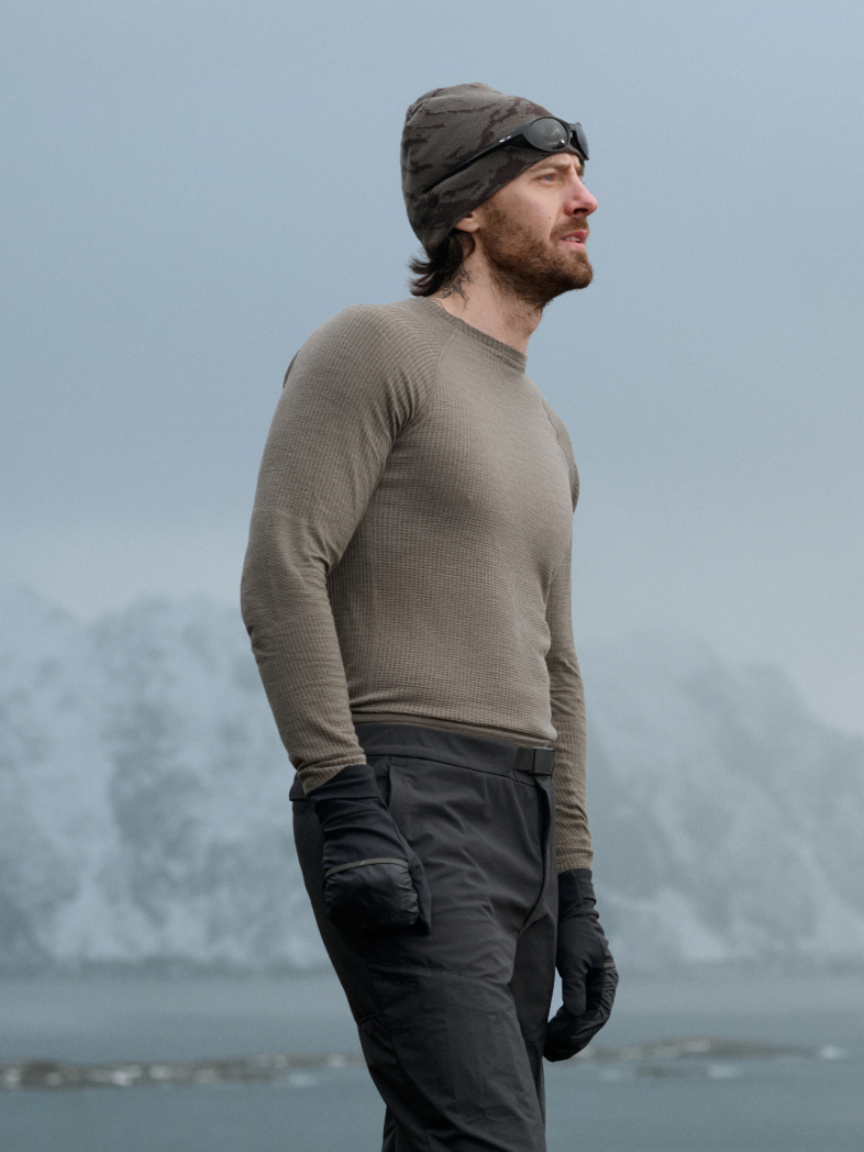 Man in olive and woman in white thermal base layers, beanies, and gloves look out at a cold, grey, mountainous landscape.