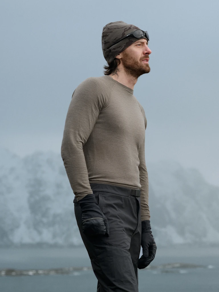 Man in olive and woman in white thermal base layers, beanies, and gloves look out at a cold, grey, mountainous landscape.