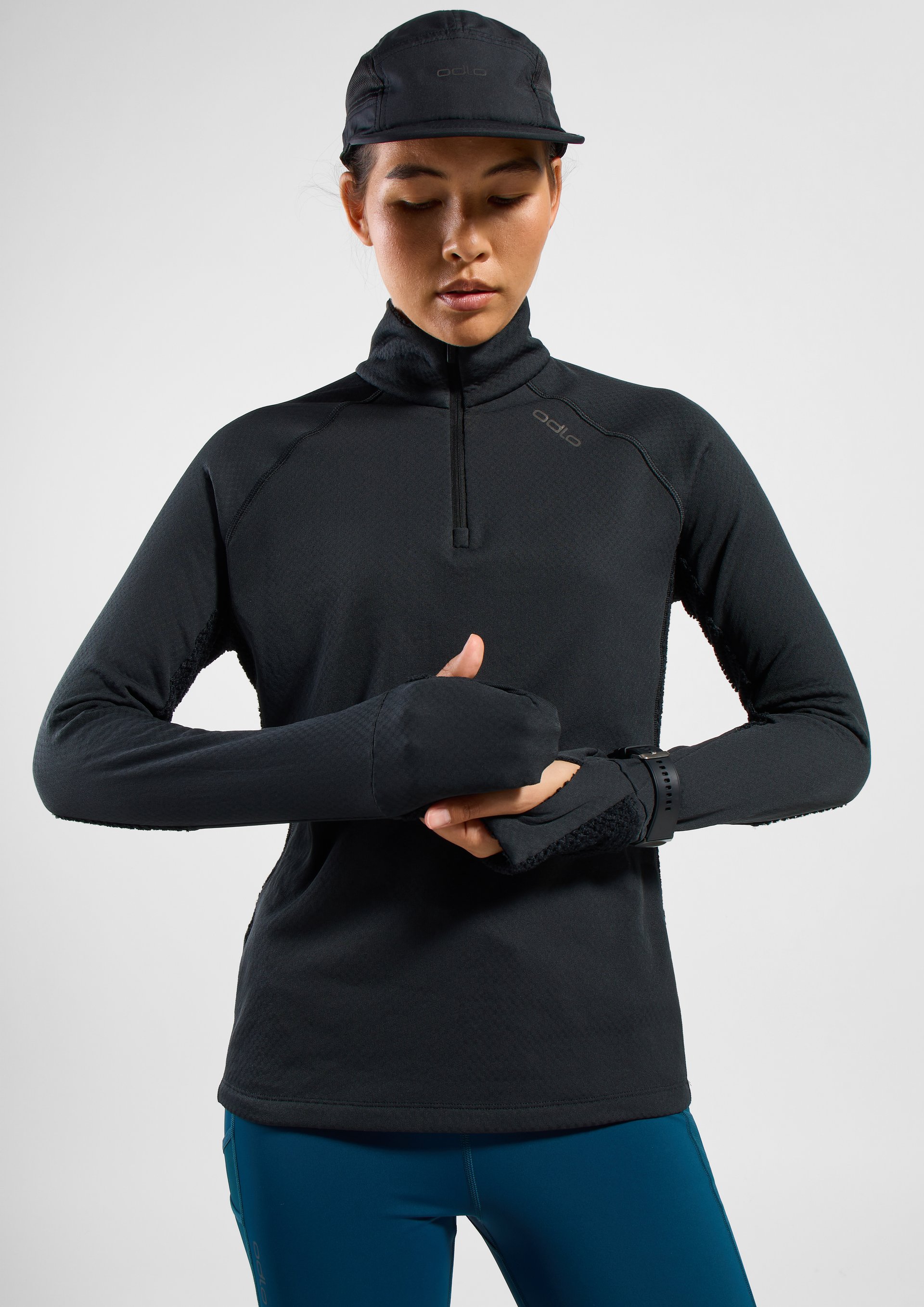 Person in a black Odlo cap and half-zip top with blue leggings, adjusting the built-in sleeve mittens over their hands.
